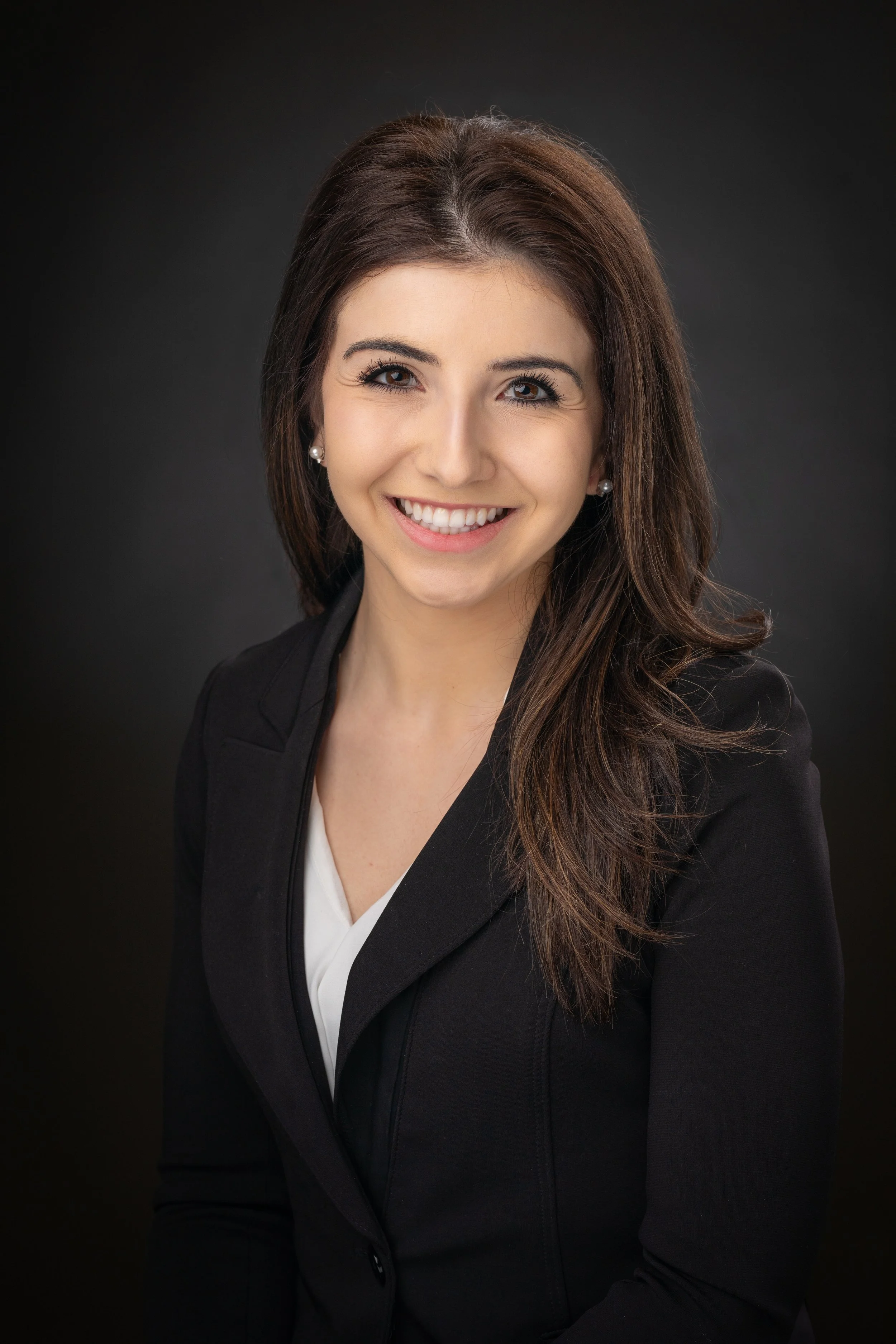 A woman with long brown hair, wearing a black blazer and white blouse, smiling against a dark background.