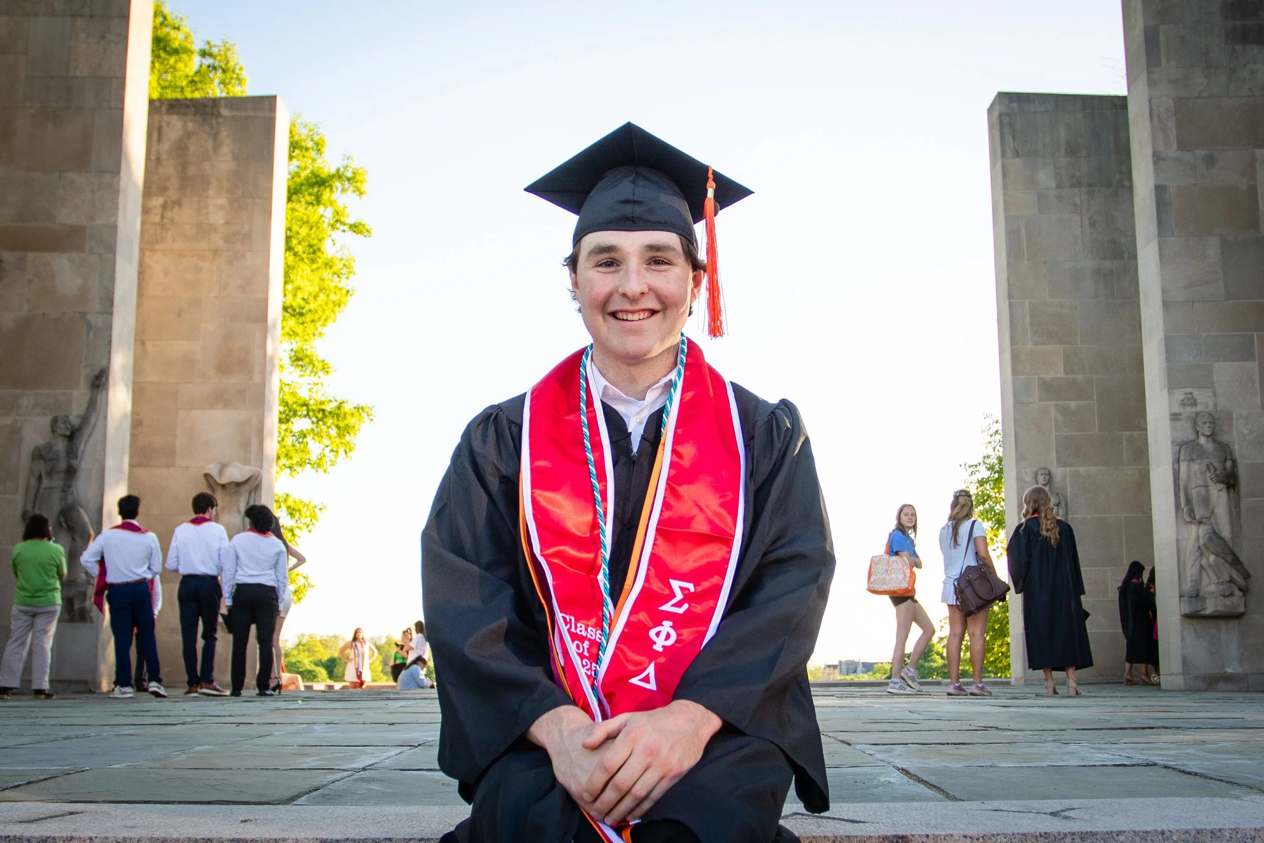 A young man in a graduation cap and gown smiling, sitting on the ground with a graduation stole and cords, at a graduation ceremony outdoors with other graduates and guests in the background, under bright sunlight and a clear sky.