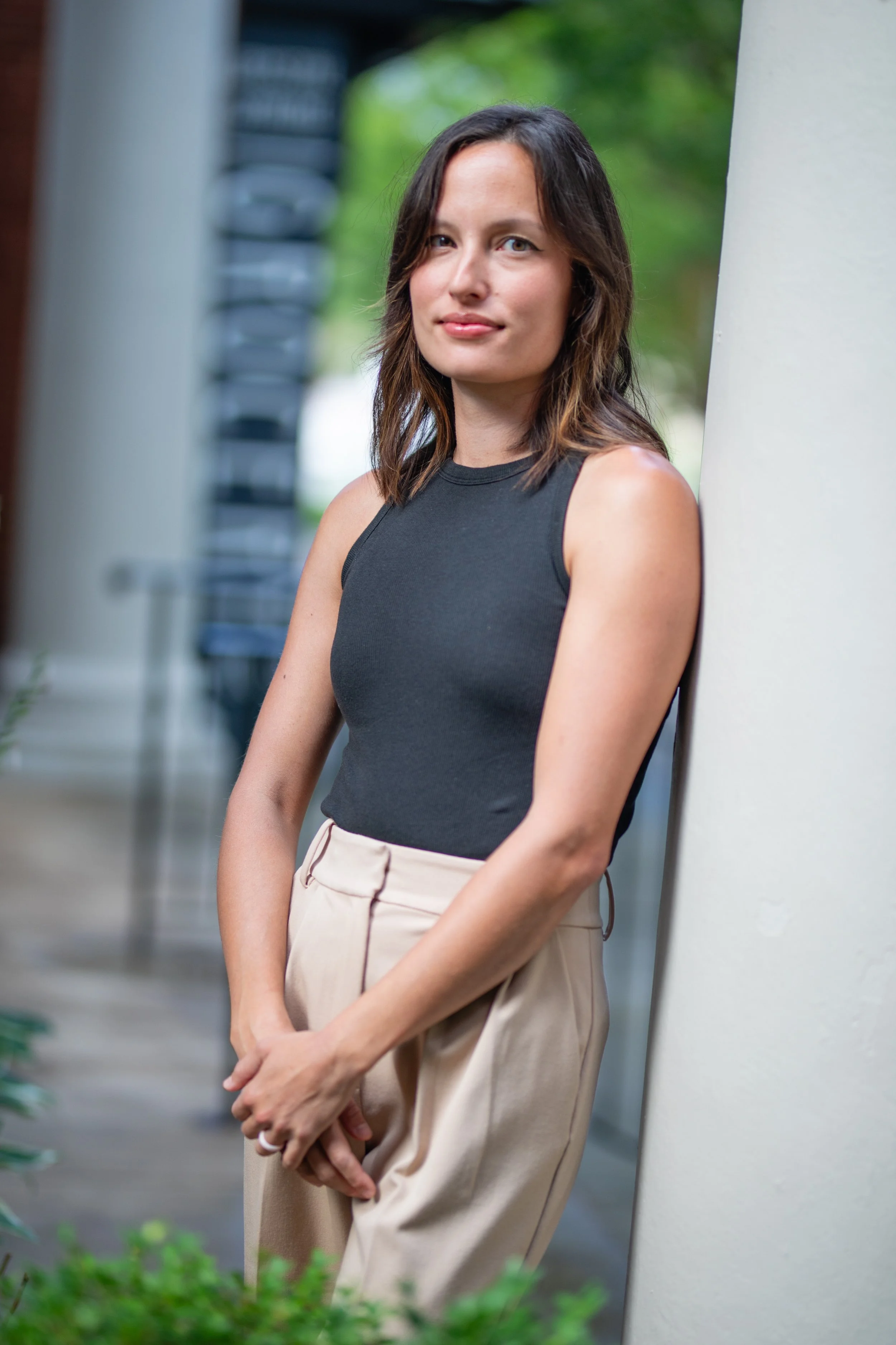 A woman leaning against a white pillar outdoors, wearing a black sleeveless top and beige pants, with trees and blurred chairs in the background.