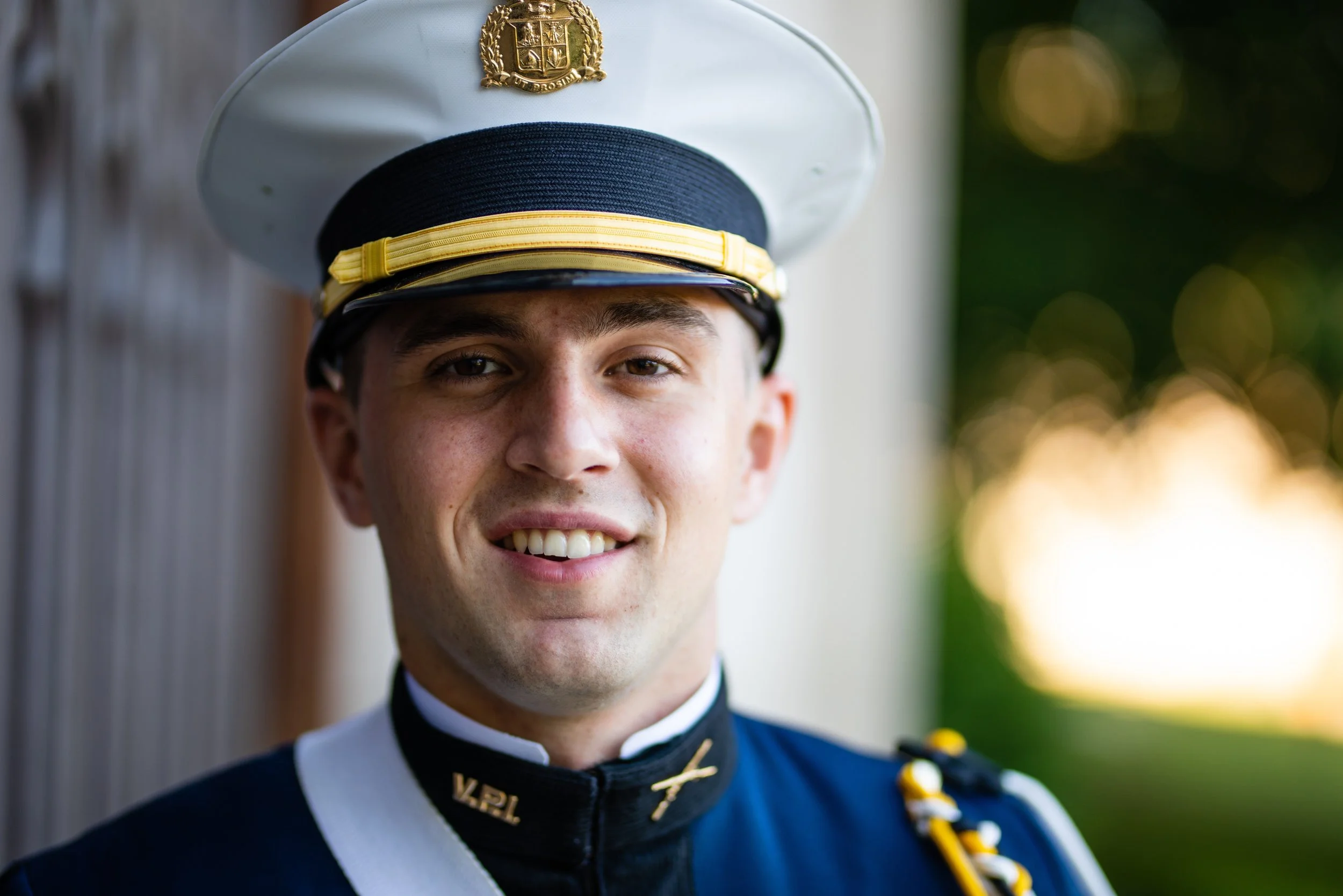 A young man in a military dress uniform, wearing a white peaked cap with a gold emblem, standing outdoors with a blurred background of greenery and sunlight.