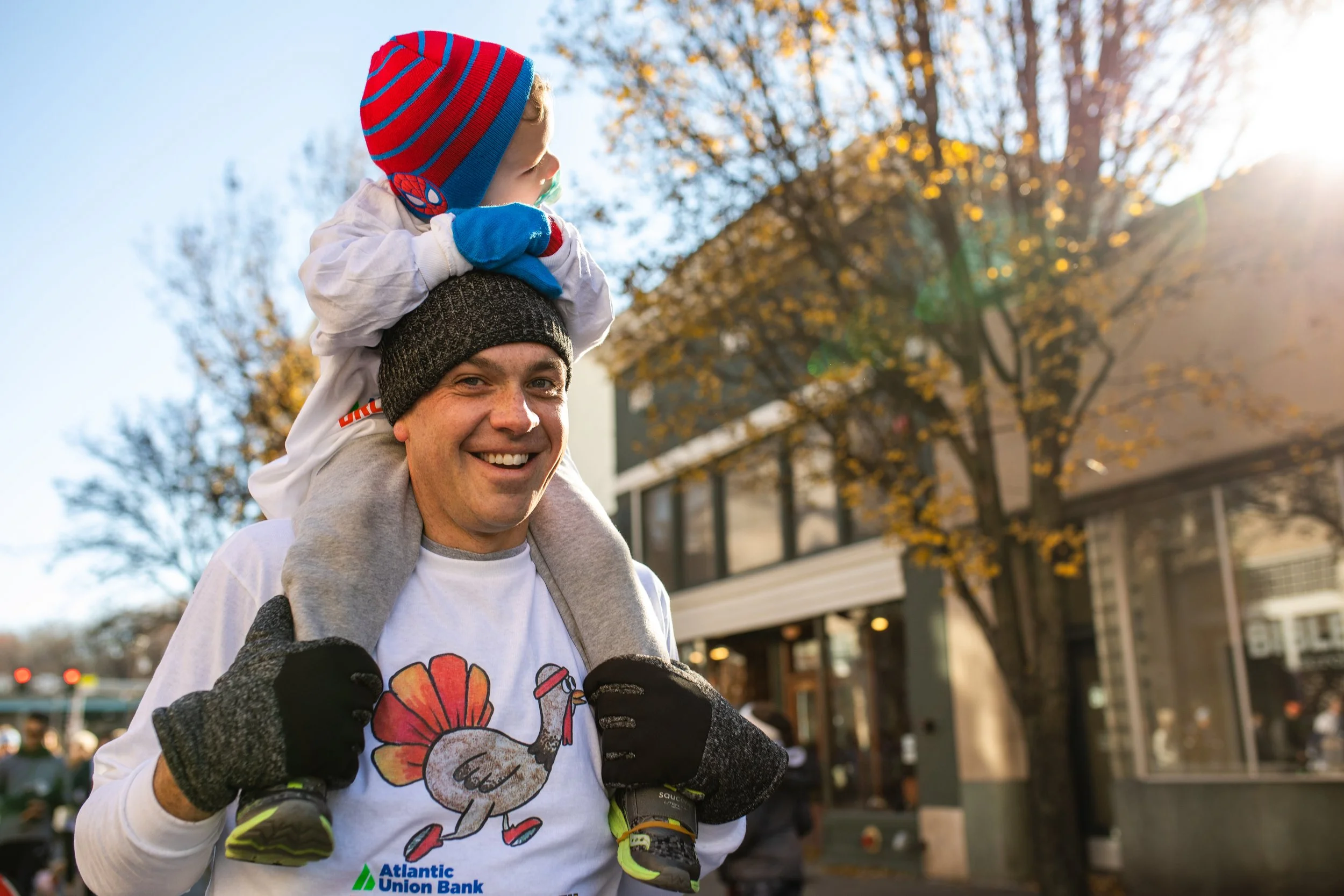 A man smiling while carrying a young child on his shoulders during a sunny day in fall, with trees and buildings in the background.