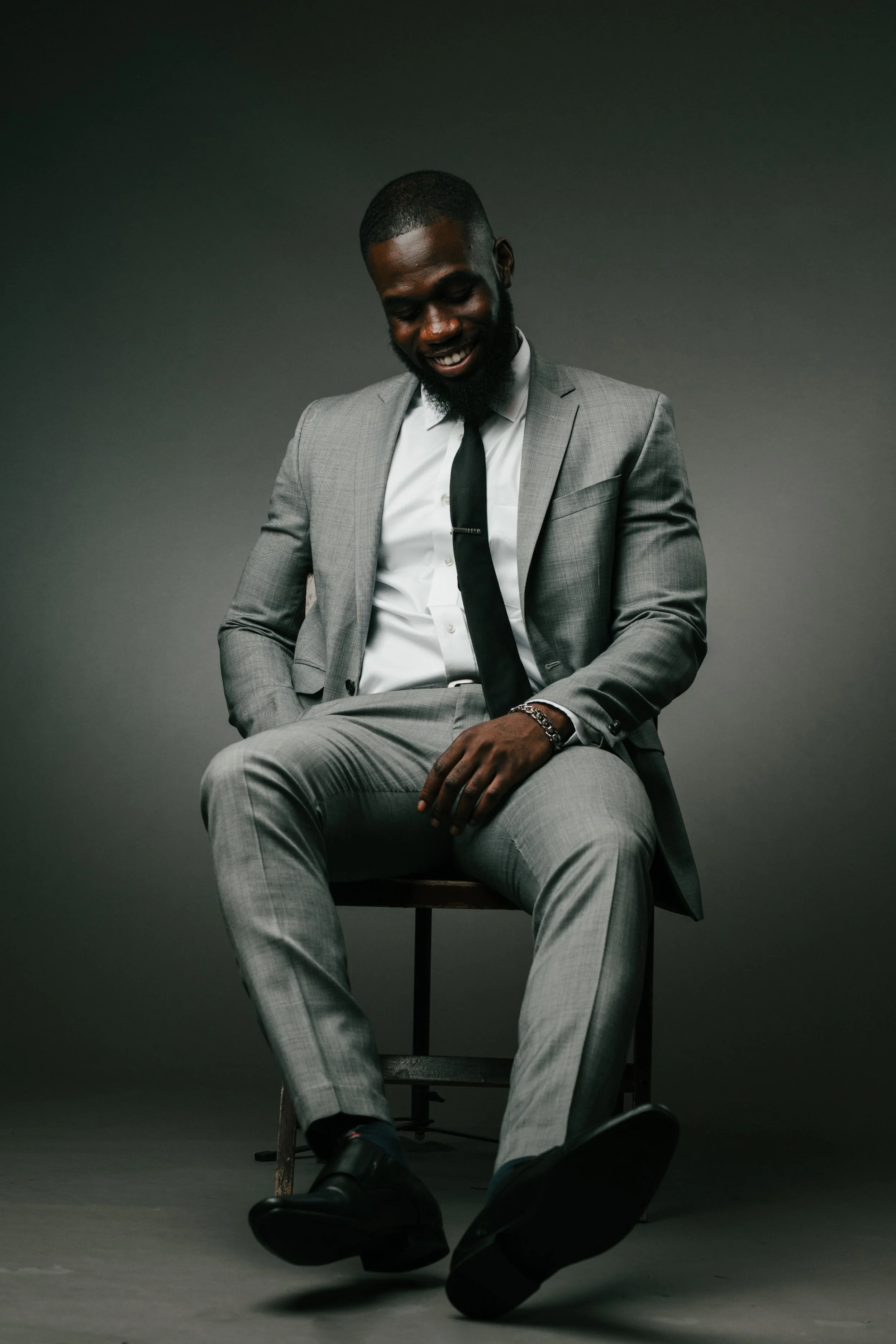 A man in a gray suit, white shirt, and black tie sitting on a wooden chair, smiling, with his head slightly tilted downward, against a dark gray background.