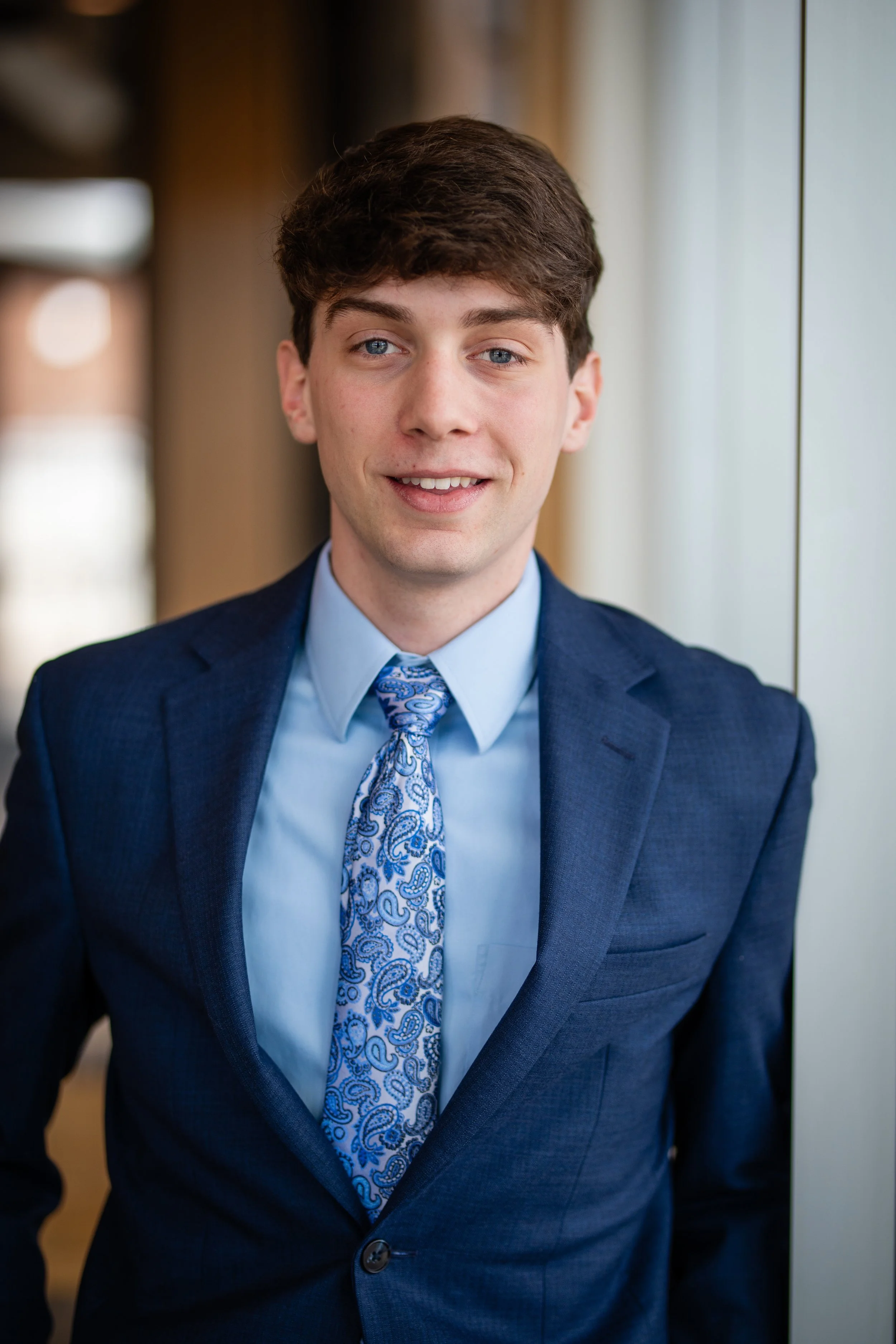 A young man with brown hair and blue eyes wearing a dark blue suit, light blue shirt, and a patterned blue tie, standing indoors near a window.