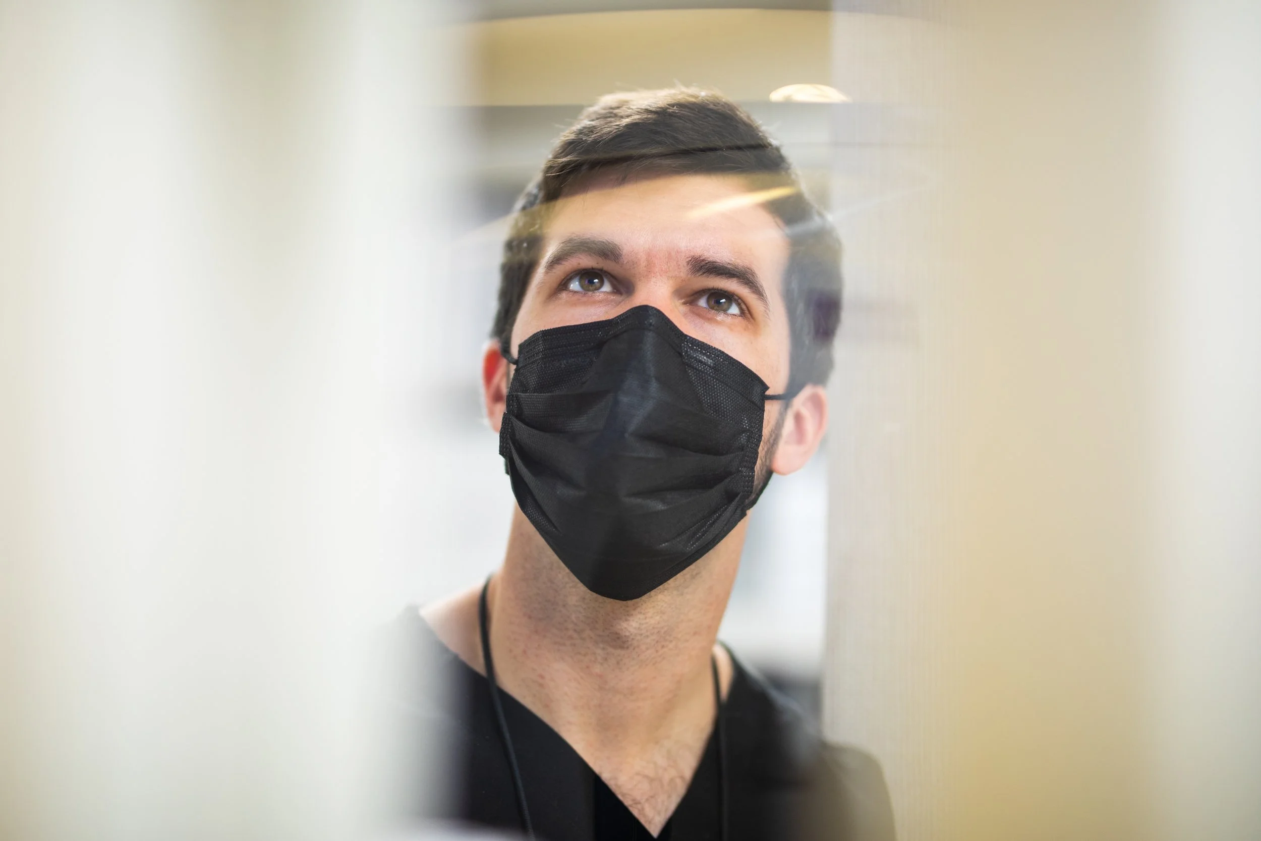 A young man with brown hair wearing a black face mask looking upward, seen through a transparent barrier in an indoor setting.