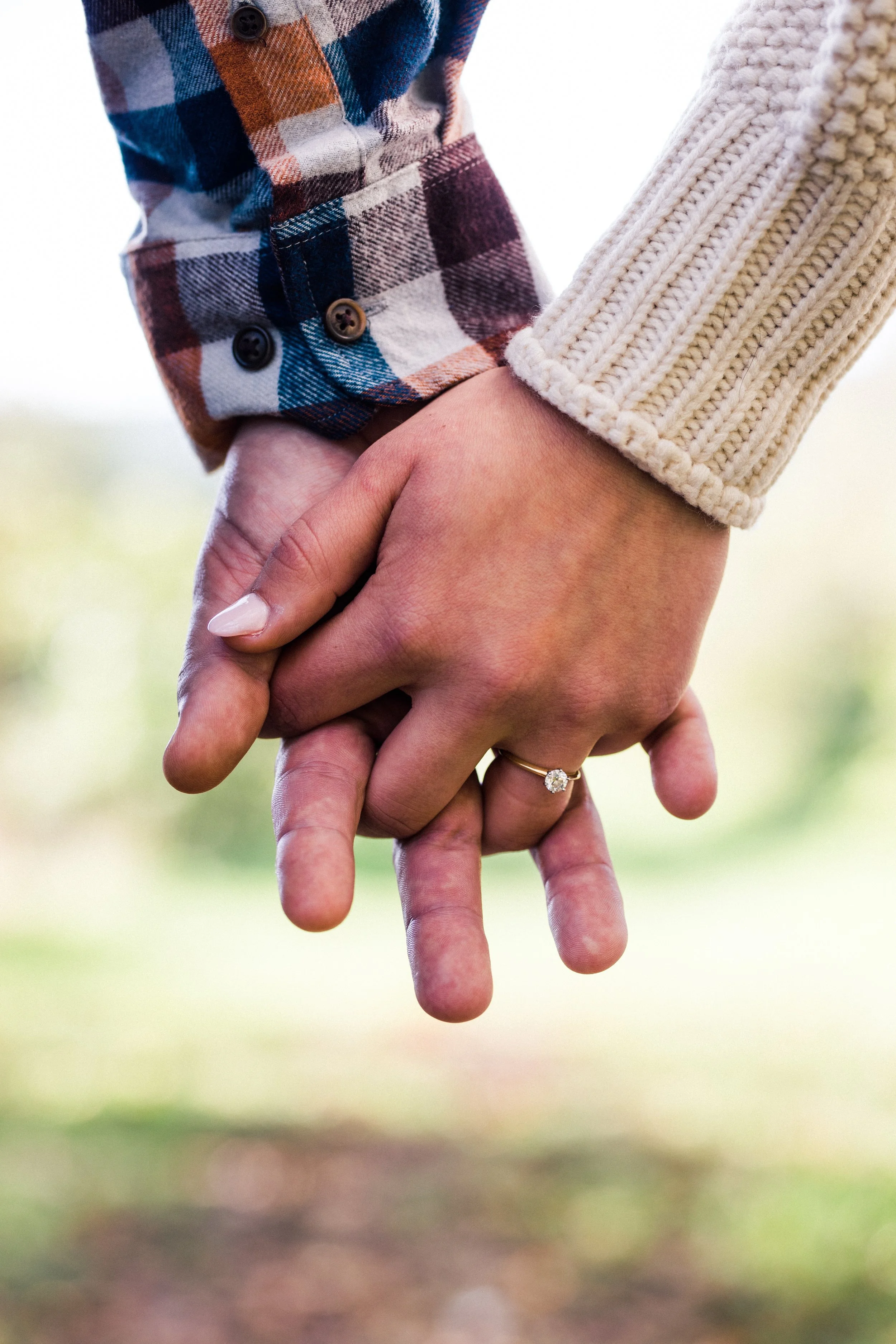 A close-up of two people holding hands, one wearing a plaid shirt and the other wearing a beige knitted sweater. The person with the plaid shirt has a ring on their finger, visible in the photo.