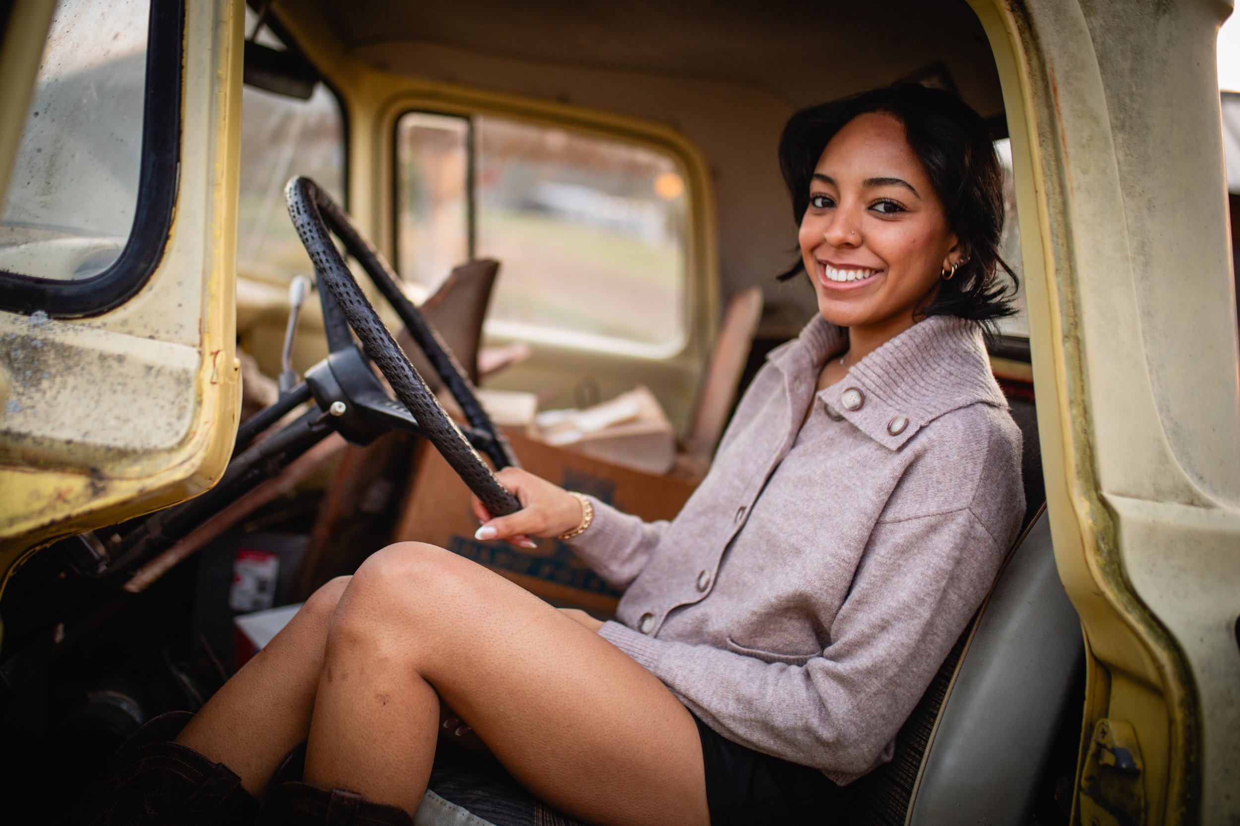 A young woman with short dark hair and a tan complexion sitting in the driver's seat of an old yellow vehicle, smiling at the camera, wearing a beige button-up sweater and shorts.