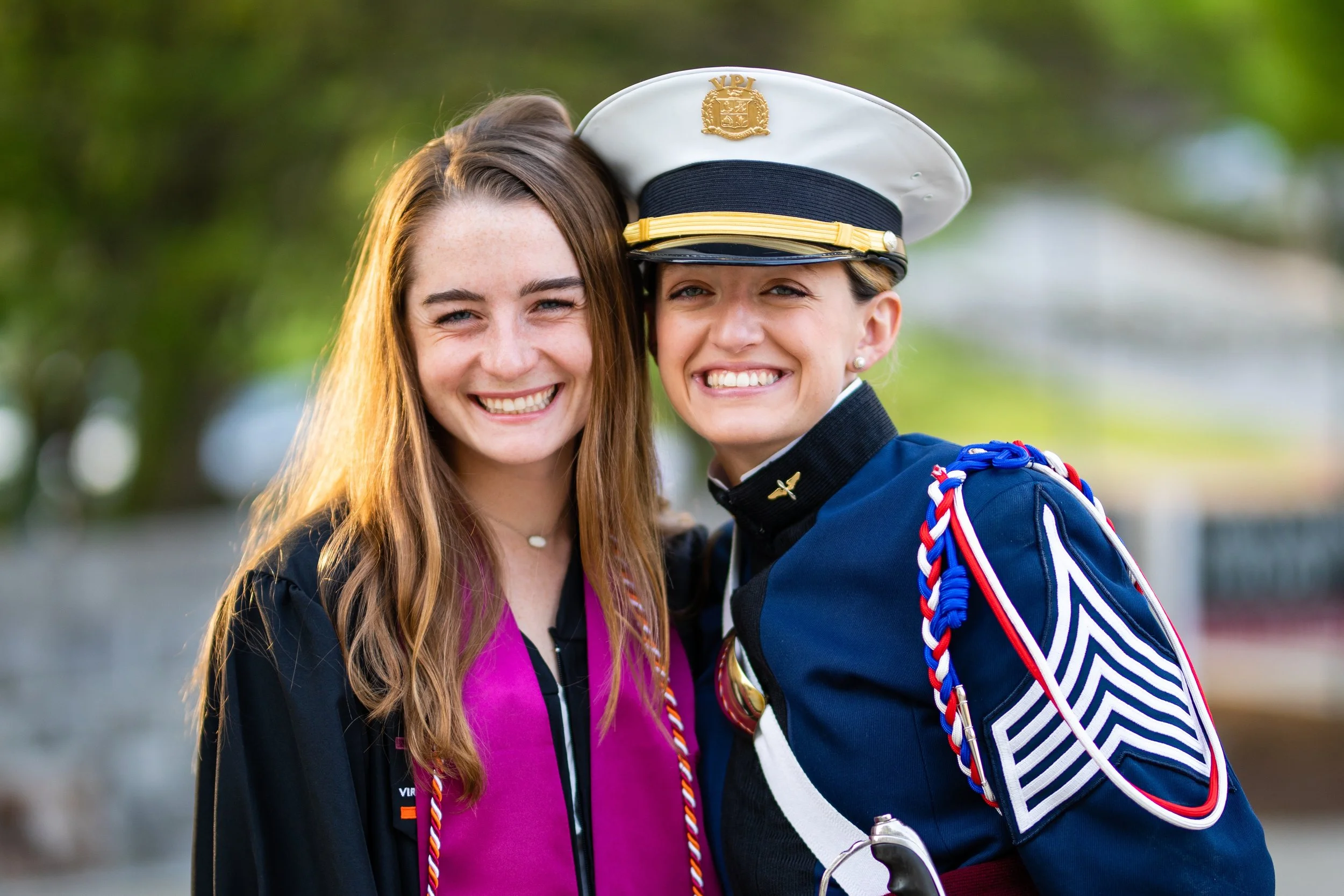 Two young women smiling, one in a graduation gown and the other in a military uniform, with a blurred outdoor background.