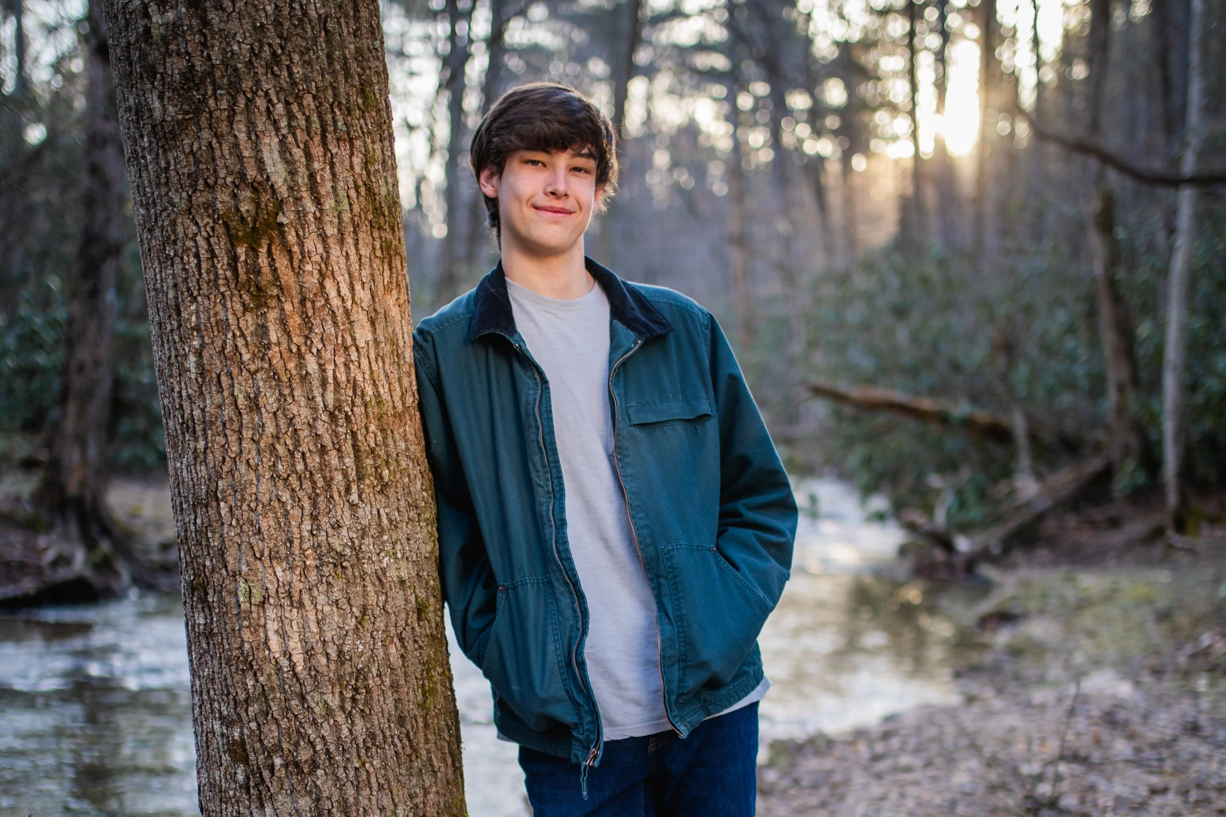 A young man with dark hair standing next to a tree with hands in pockets, smiling, in a forest near a small creek at sunset.