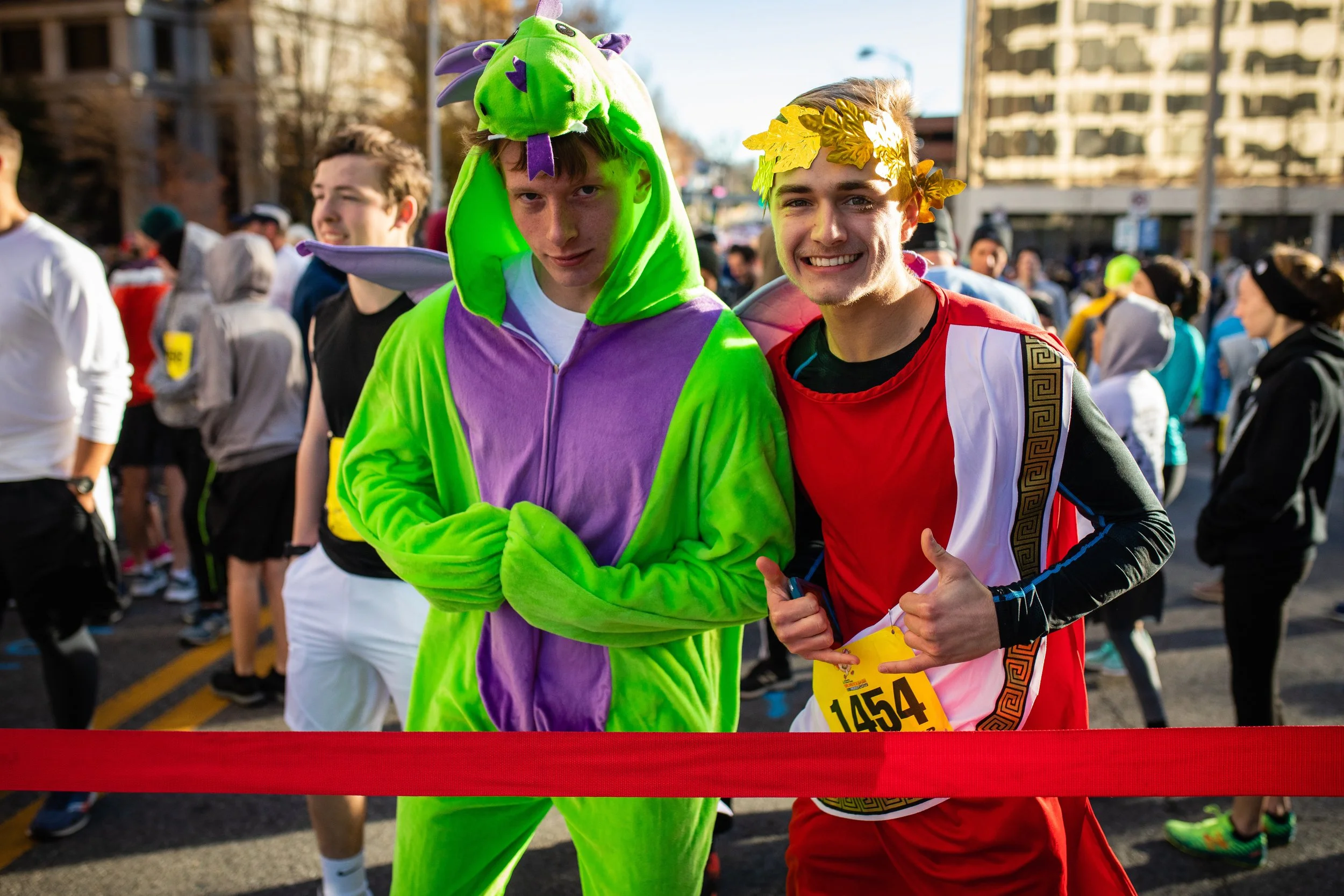 Two young men at a race event, dressed in colorful costumes. One is in a bright green and purple dinosaur costume, and the other is in an ancient Greek or Roman-inspired outfit with a laurel wreath on his head, smiling and making a shaka sign. There 
