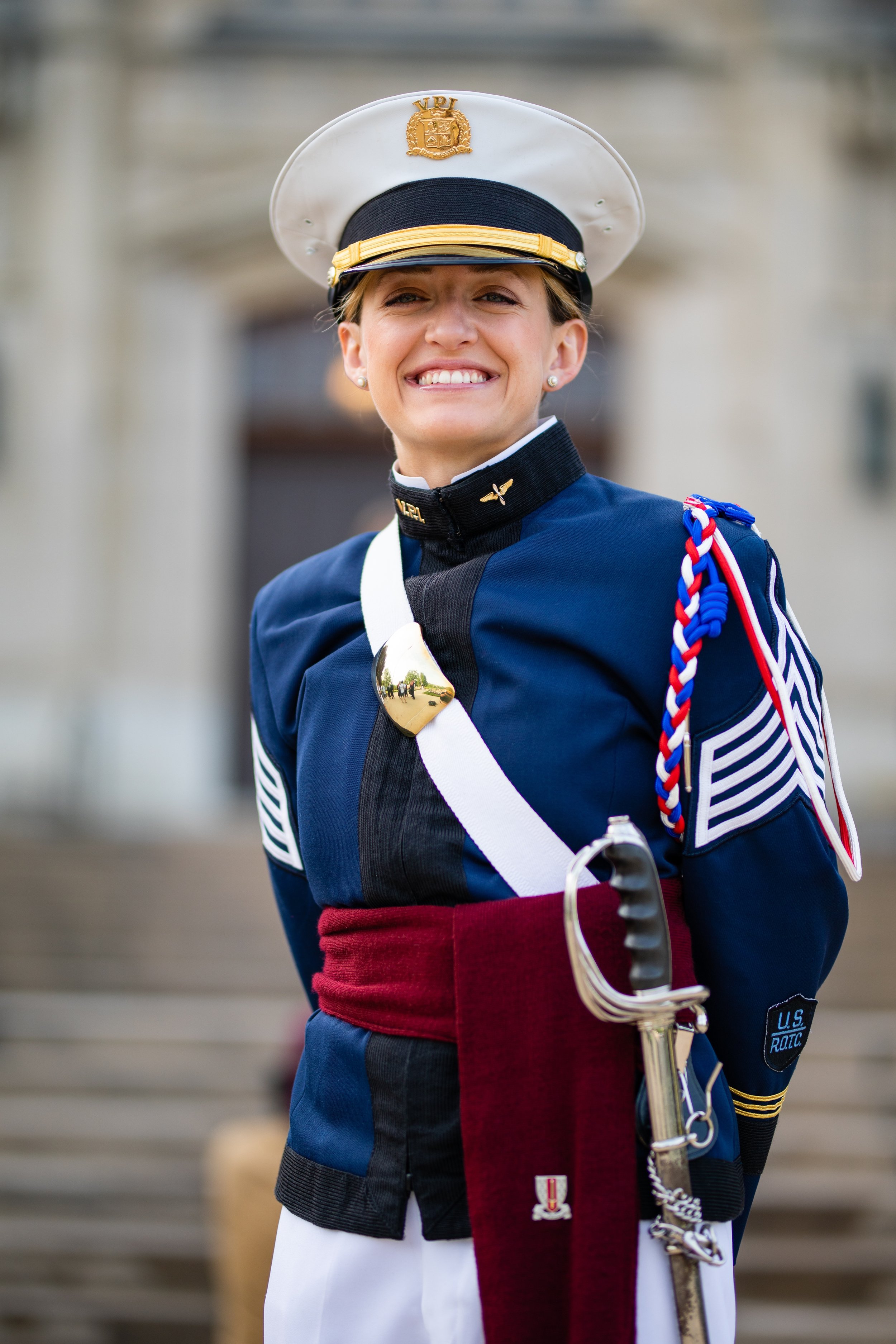 A smiling woman dressed in a U.S. military uniform, including a blue jacket with chevrons, a white peaked cap with gold emblem, and a sash with a sword.