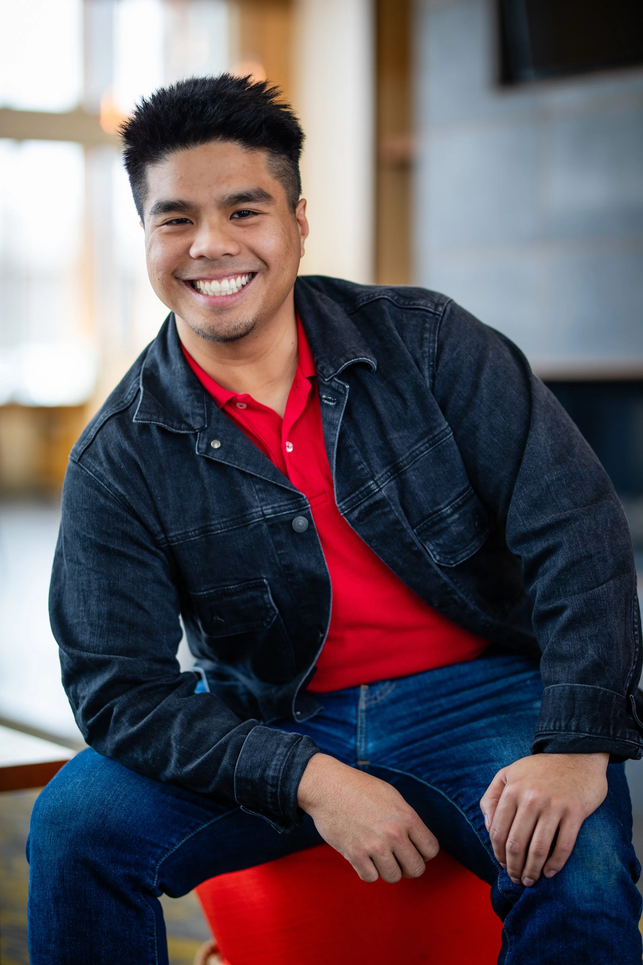 A smiling man with black hair, wearing a black denim jacket over a red shirt, sitting on a red stool indoors with blurred background.