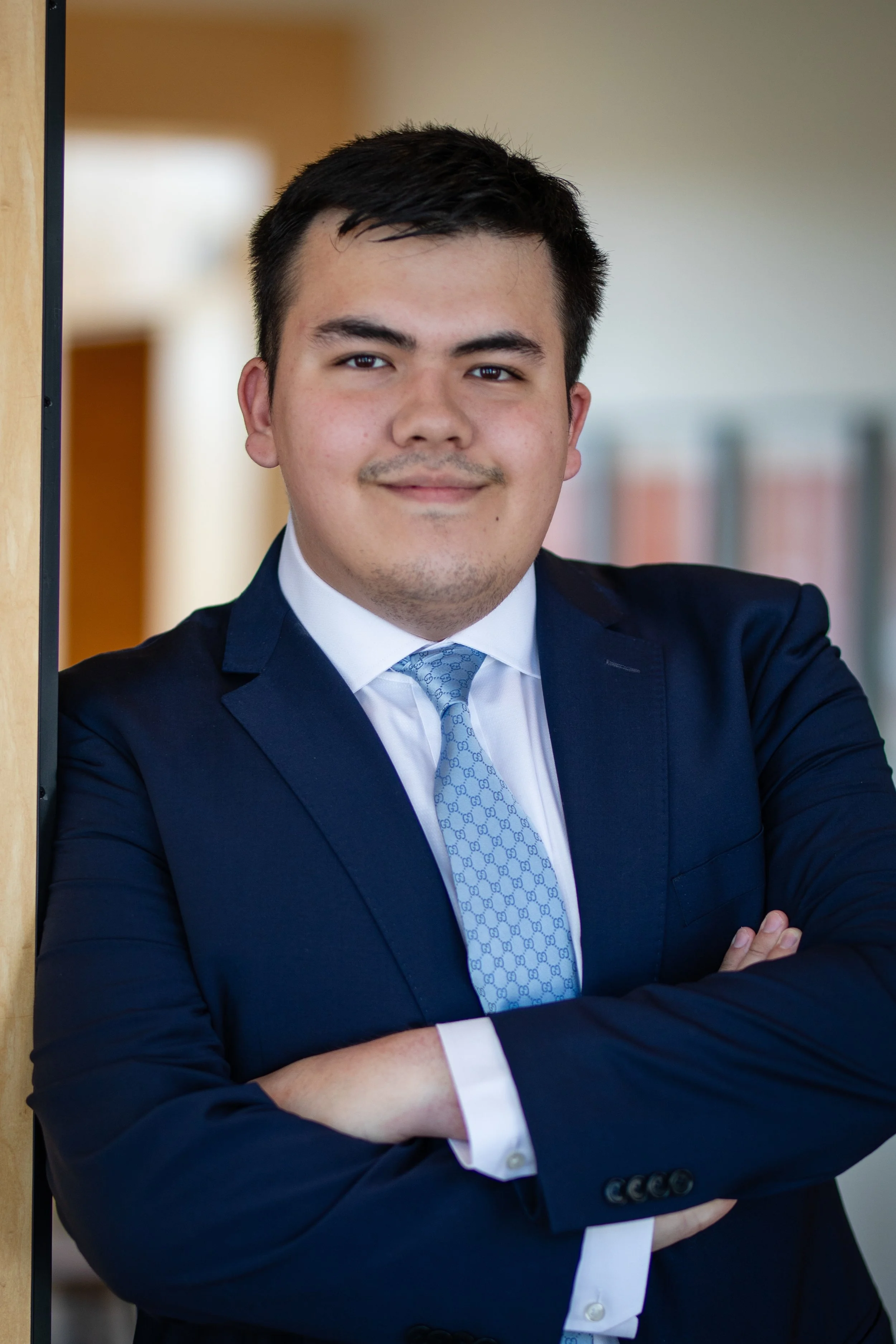 A young man in a dark blue suit and light blue tie standing with arms crossed, smiling, in an indoor setting.