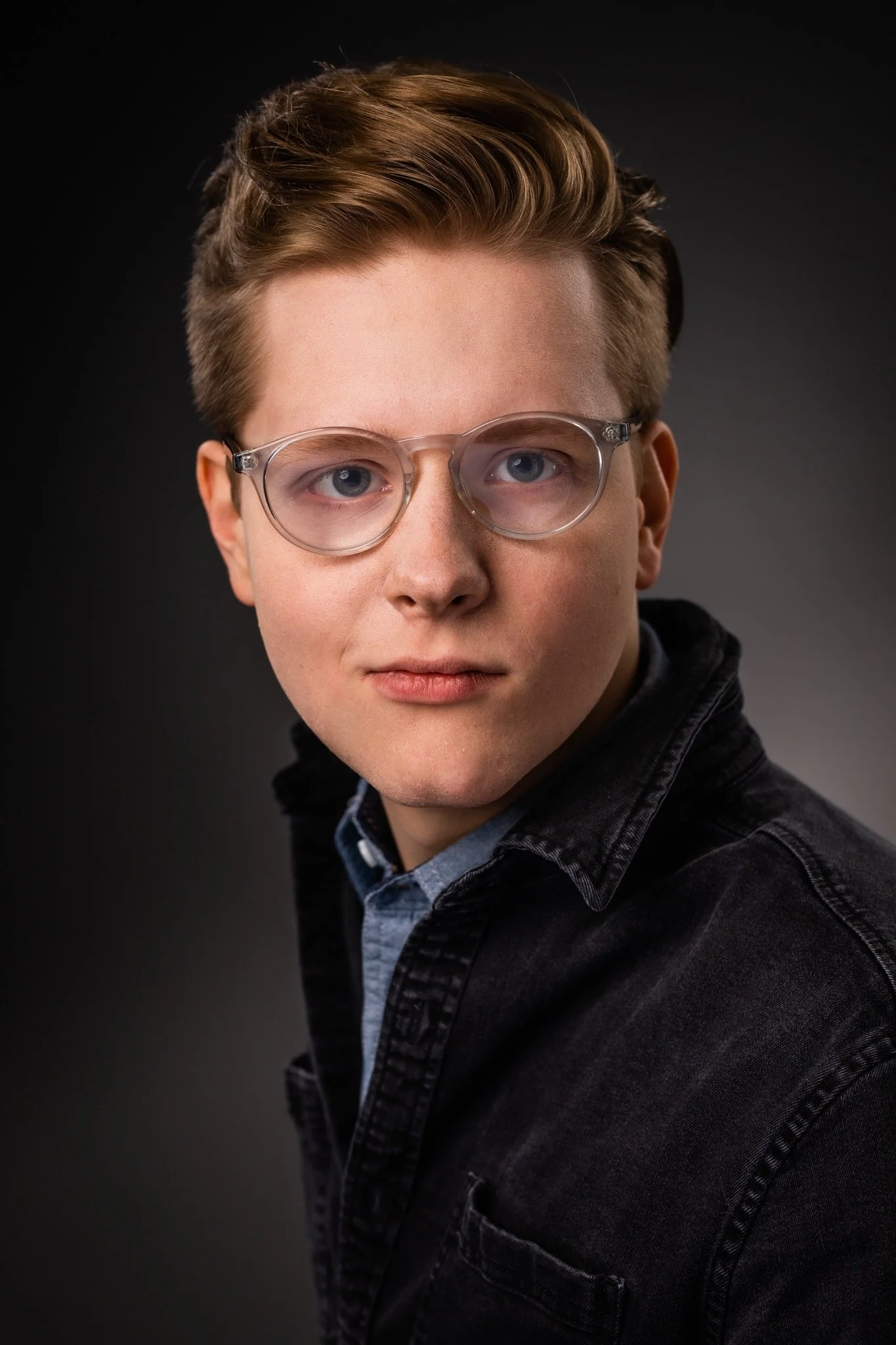 Portrait of a young man with blond hair, blue eyes, wearing clear glasses and a dark denim jacket, against a dark background.
