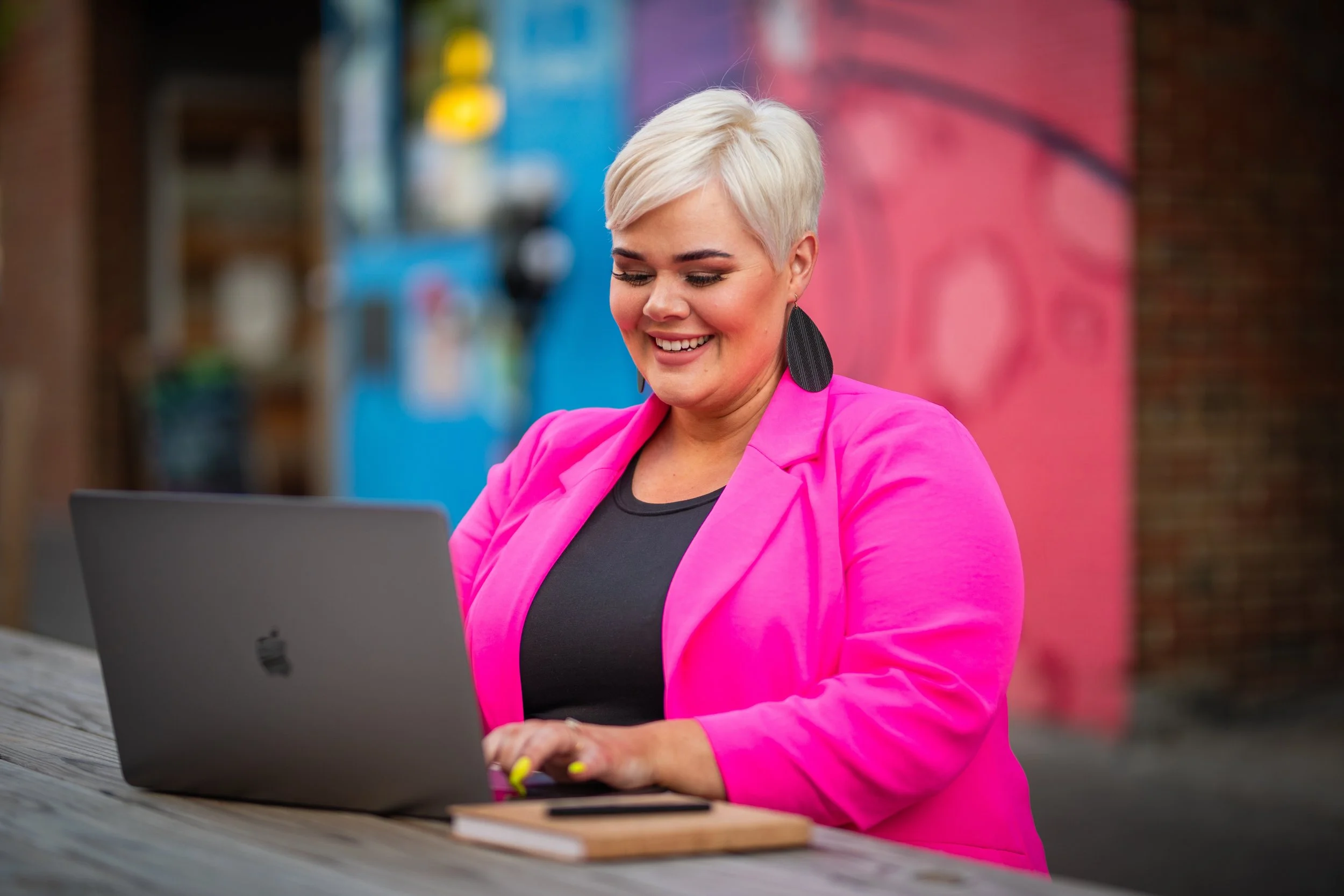 A woman with short platinum blonde hair wearing a bright pink blazer and black top, sitting outdoors at a wooden table, smiling as she works on a silver MacBook laptop.