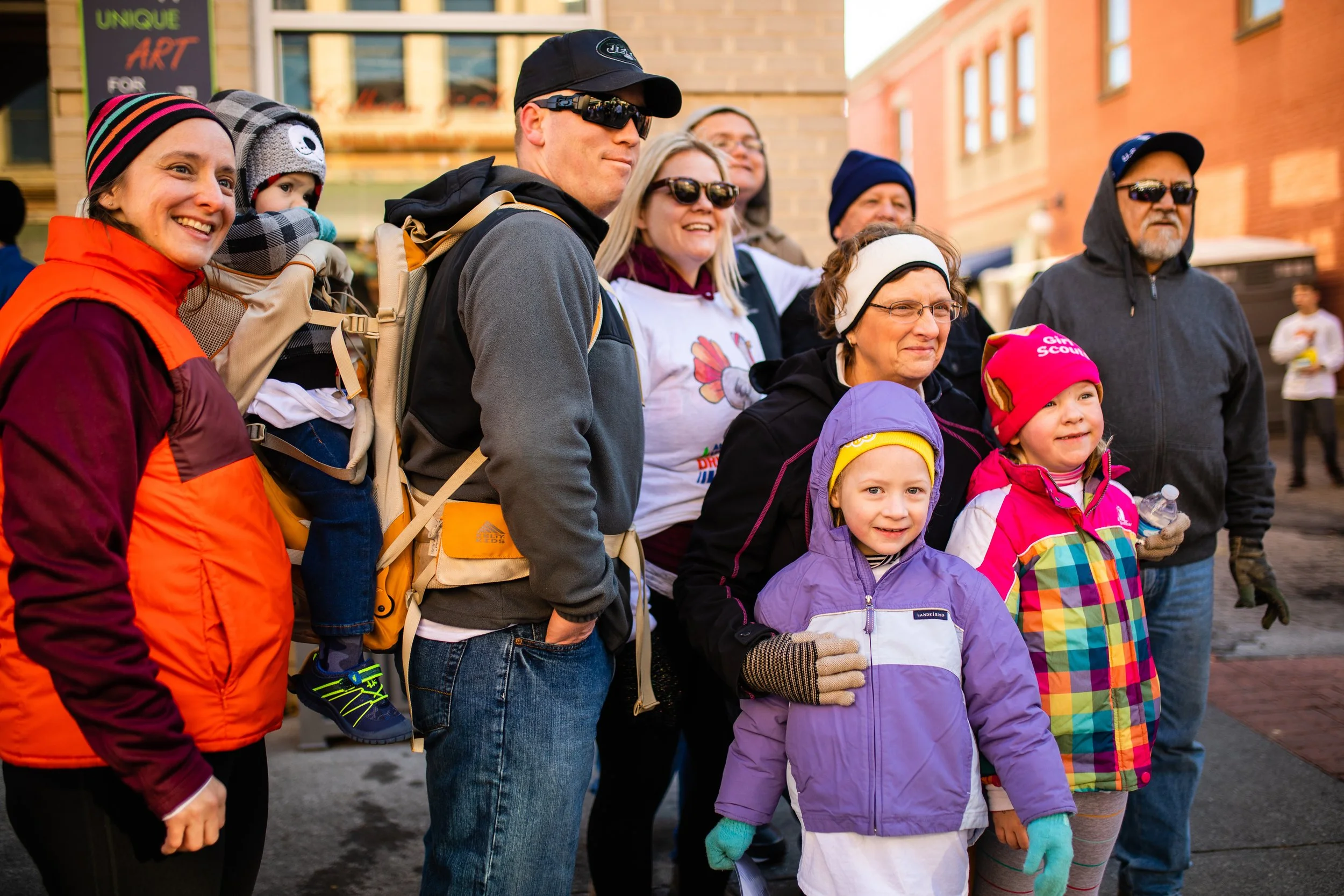 A group of people, including children and adults, outdoors in a city setting, wearing colorful jackets and winter accessories. They are smiling and posing for a photo.