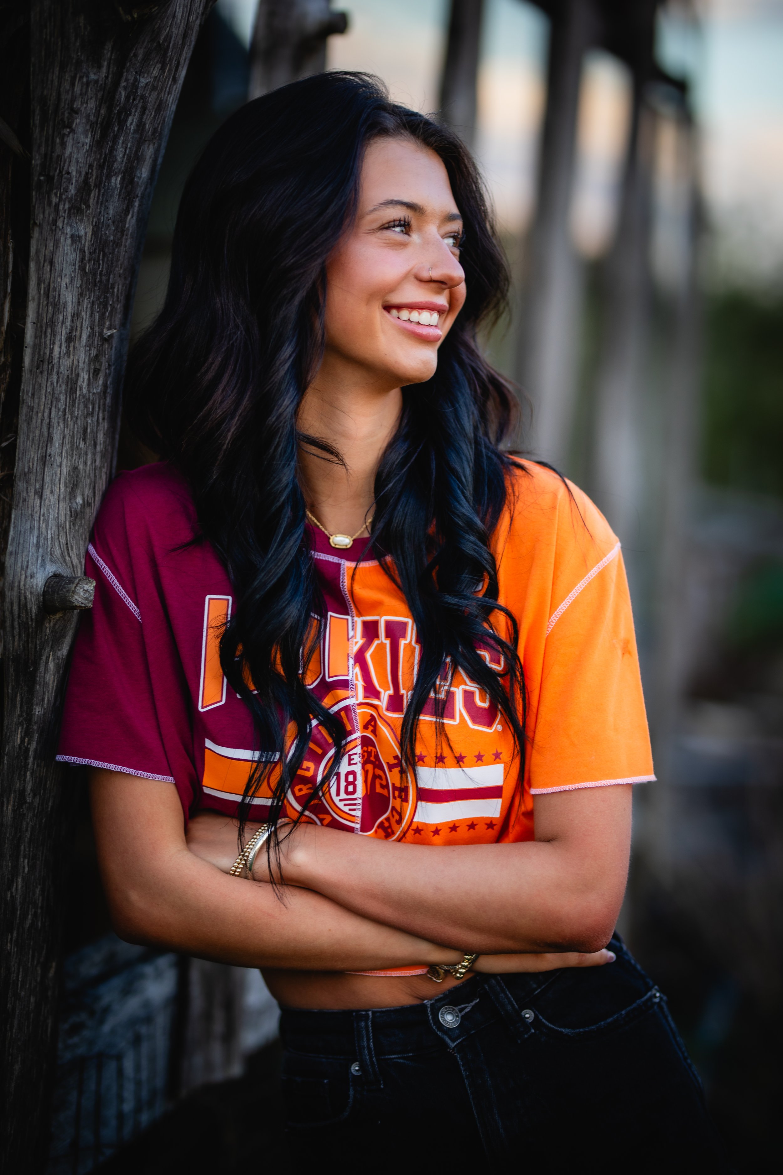 A young woman with long dark hair with blue highlights smiling and leaning against a wooden fence at sunset, wearing a colorful sports jersey.