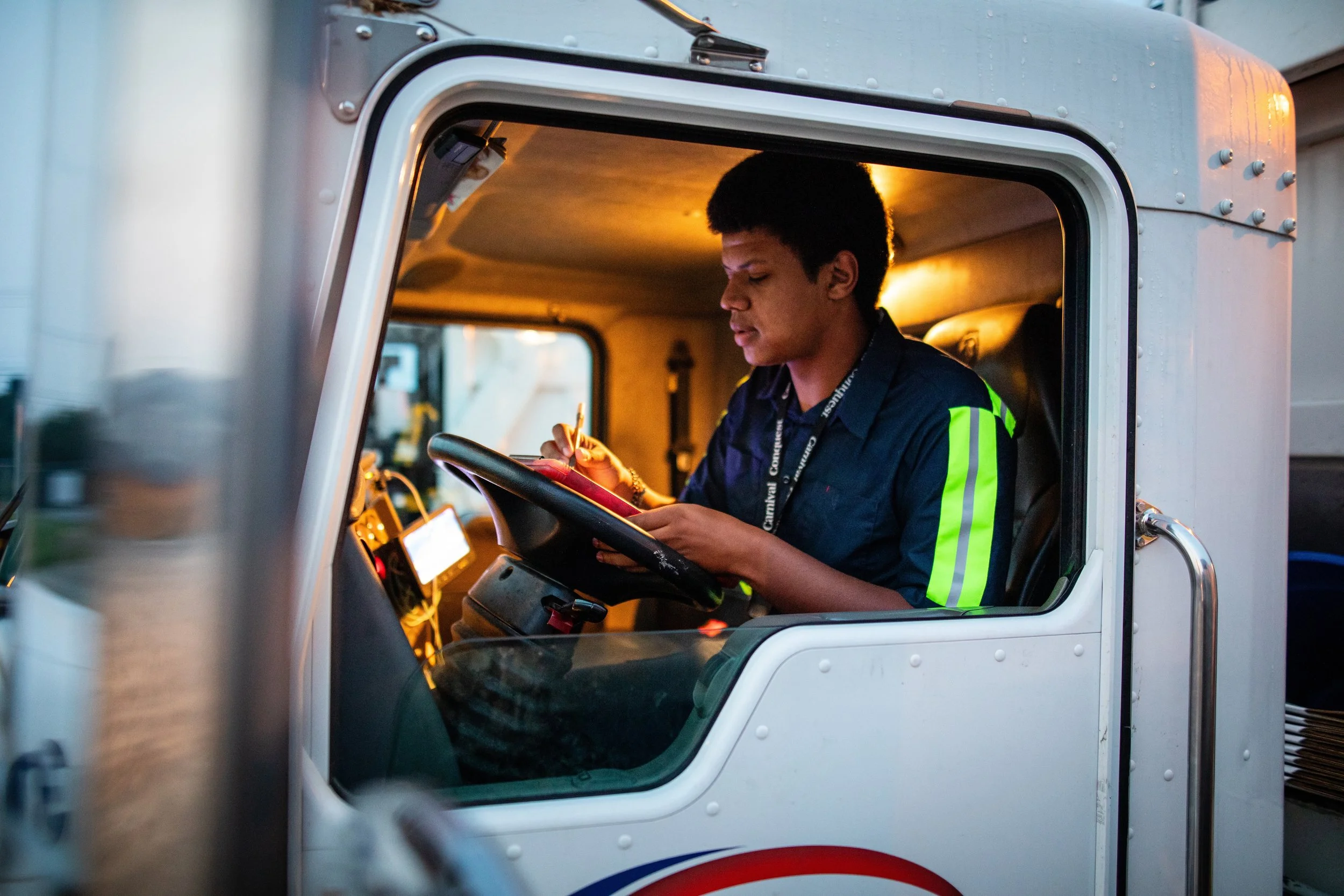 A young male truck driver sitting inside the cab of a large truck, writing on a notepad. The reflection of warm interior lights is visible on the window, and he is wearing a dark blue shirt with neon green reflective stripes.