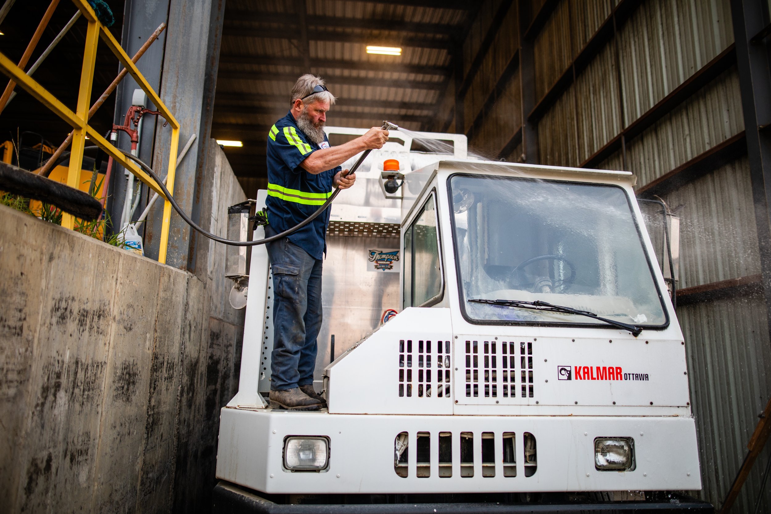A man with a beard wearing a blue uniform and safety glasses is standing on a small industrial vehicle inside a warehouse. He is holding a hose and spraying something, with the vehicle labeled "Kalmar Ottawa." The warehouse has metal walls and indust