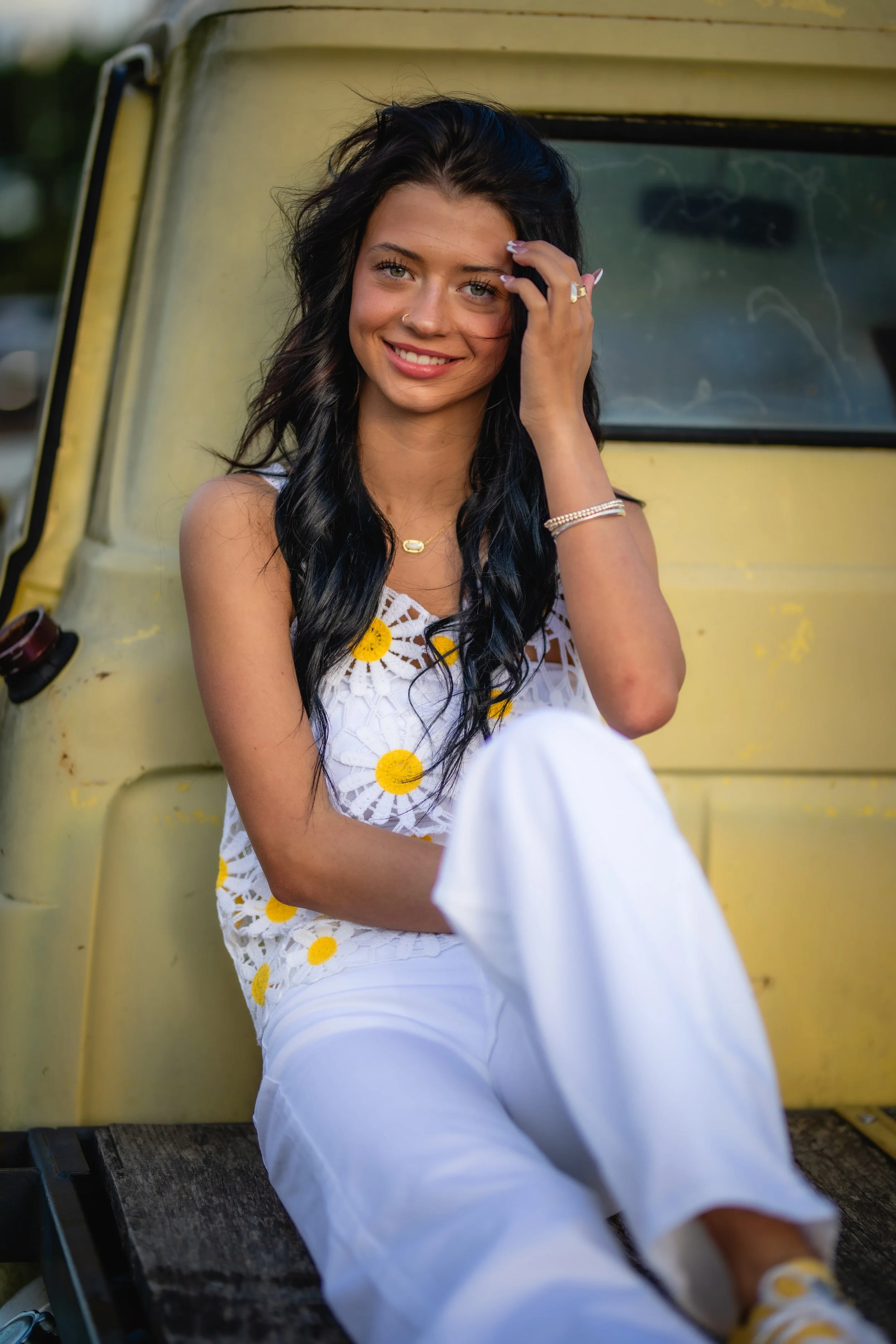A young woman with long dark hair, smiling and sitting in front of a yellow vehicle, wearing a white dress with yellow daisies.