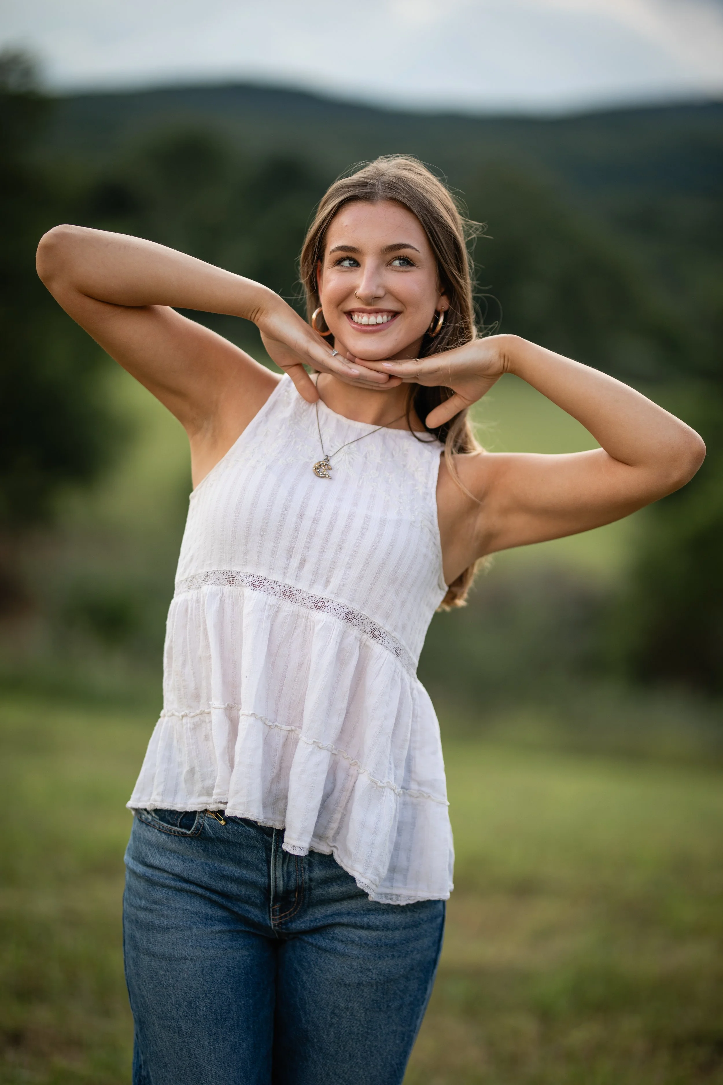 A young woman smiling outdoors with a scenic mountain background, wearing a white sleeveless top and blue jeans.