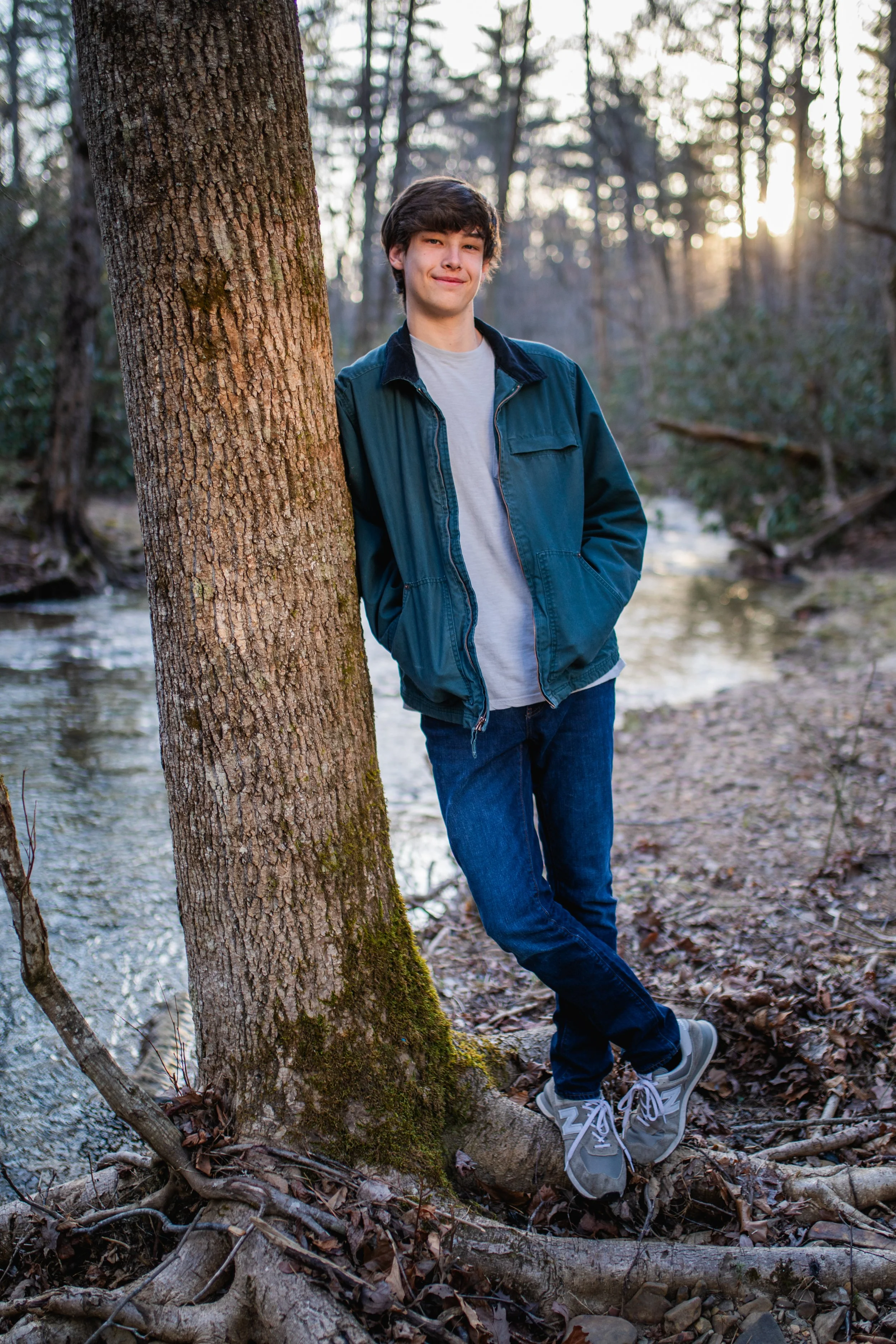 A teenage boy standing outdoors in a wooded area by a creek, leaning against a tree with one foot crossed over the other, during sunset.