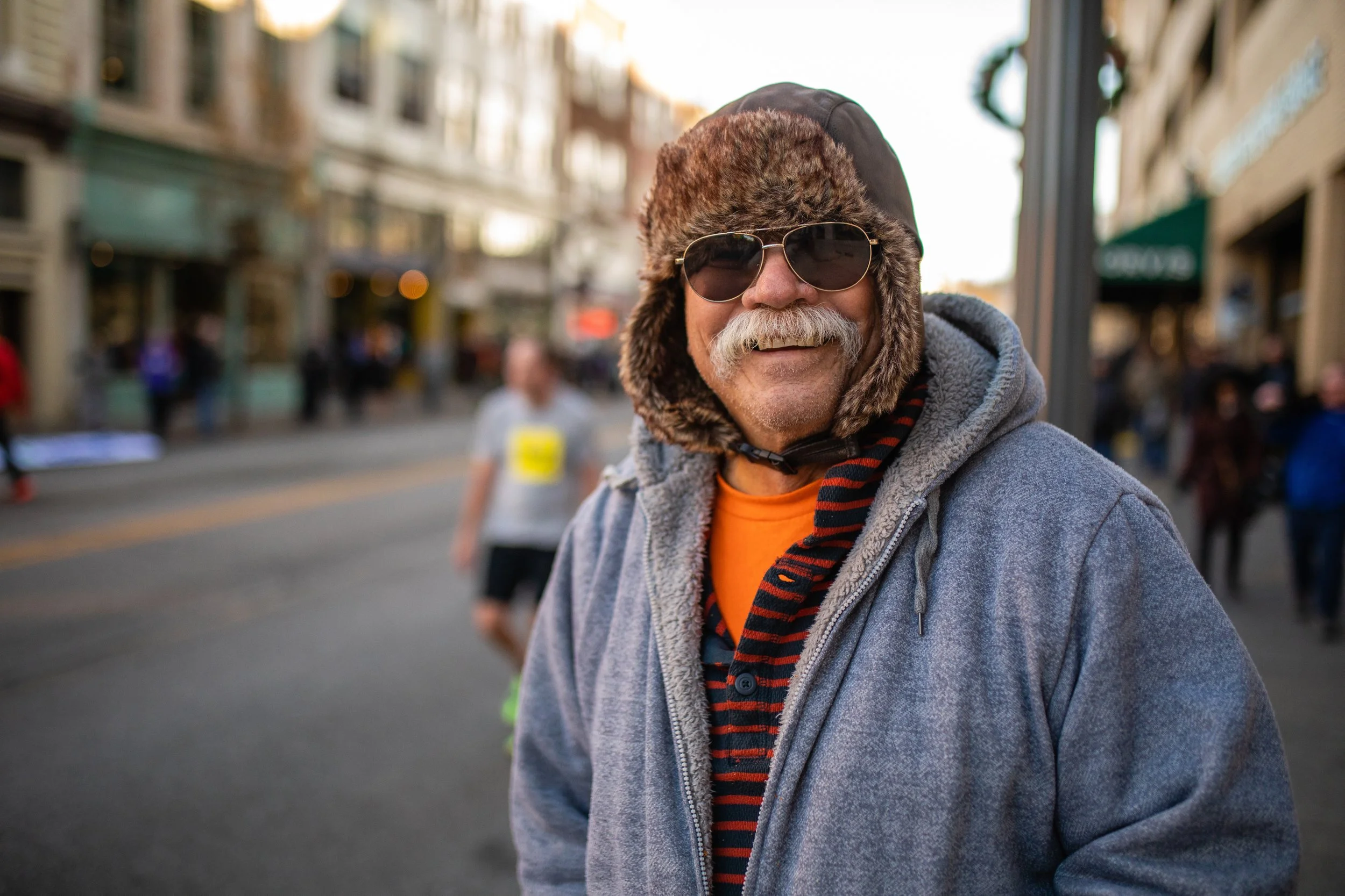 A smiling man with a mustache wearing aviator sunglasses, a fur-trimmed hat, a gray hoodie, and a striped scarf, standing on a city street during daylight.