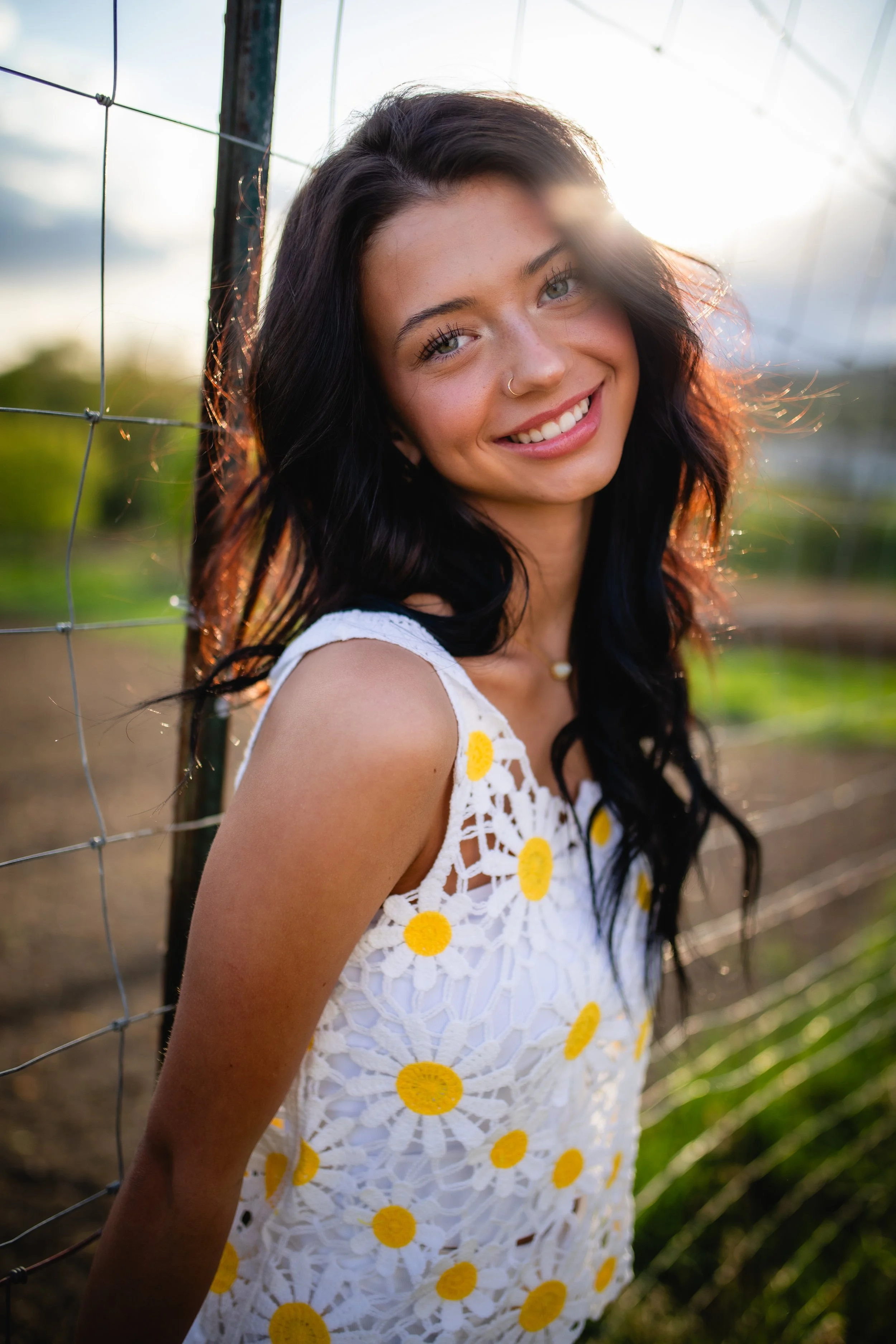 A young woman with dark hair and a nose piercing smiling outdoors at sunset, wearing a white crocheted dress with yellow floral patterns, leaning against a fence.