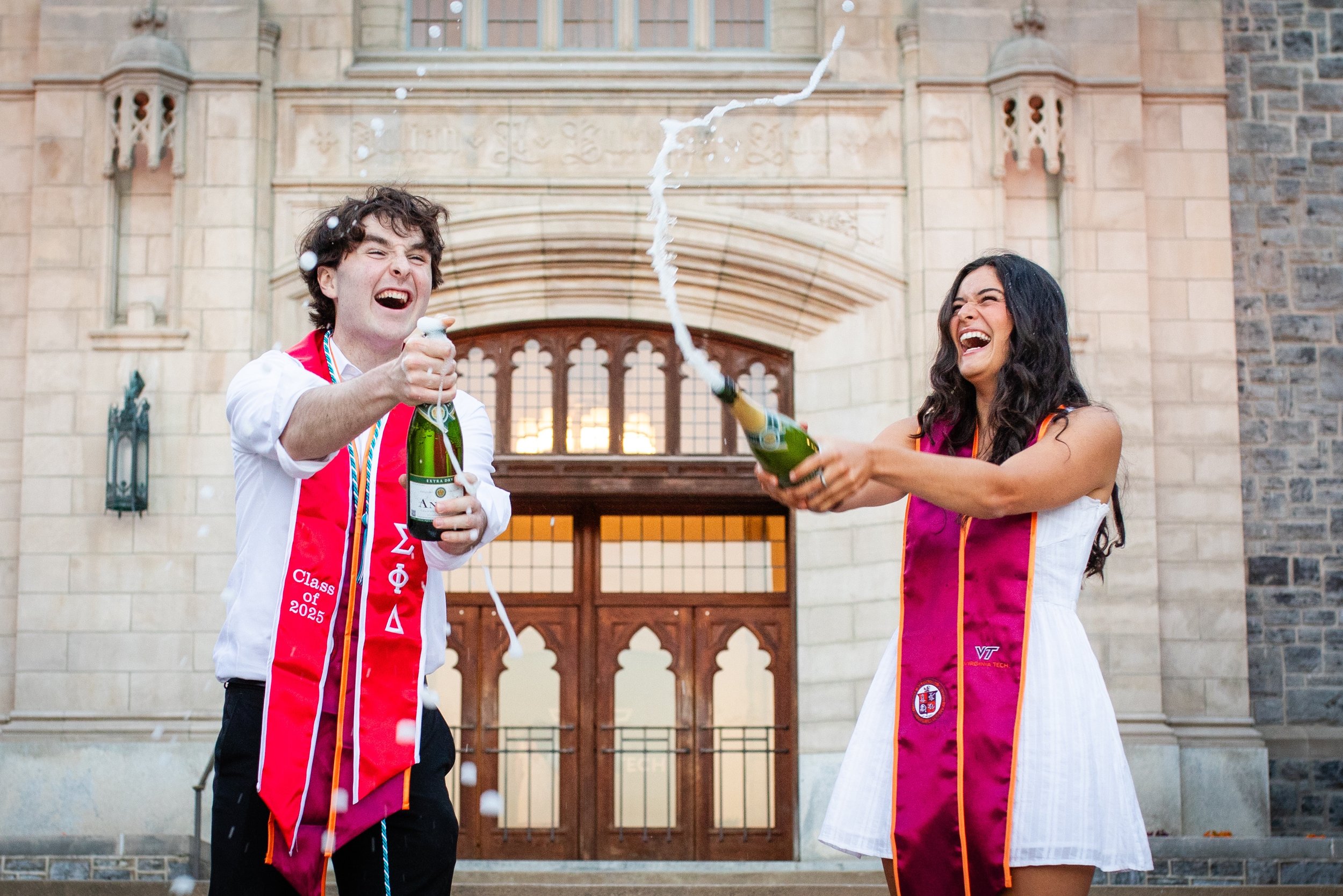 Two young adults celebrating graduation outside a historic building, popping champagne and laughing, wearing graduation stoles.