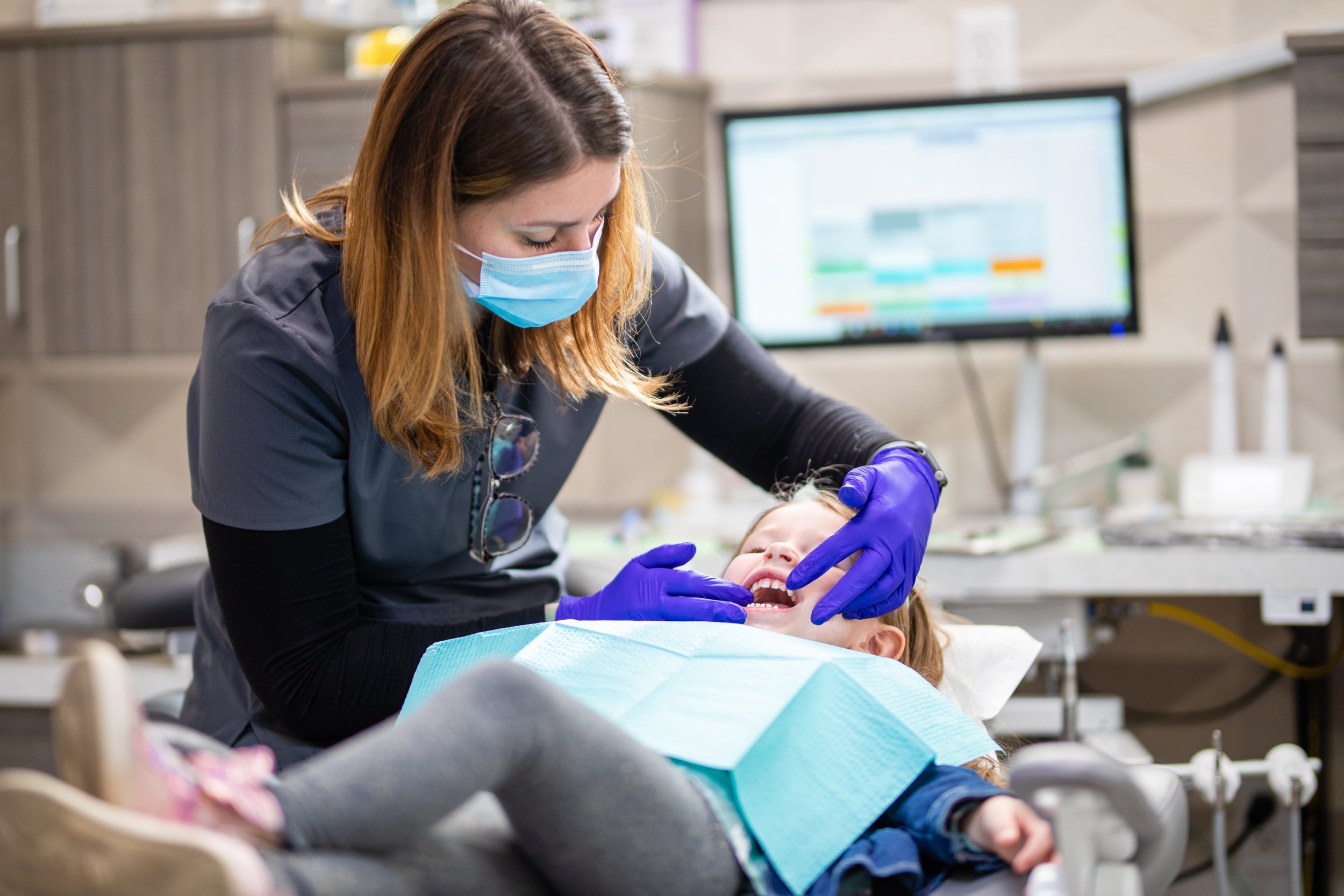 Dentist wearing a face mask and gloves examining a young girl patient at a dental clinic.