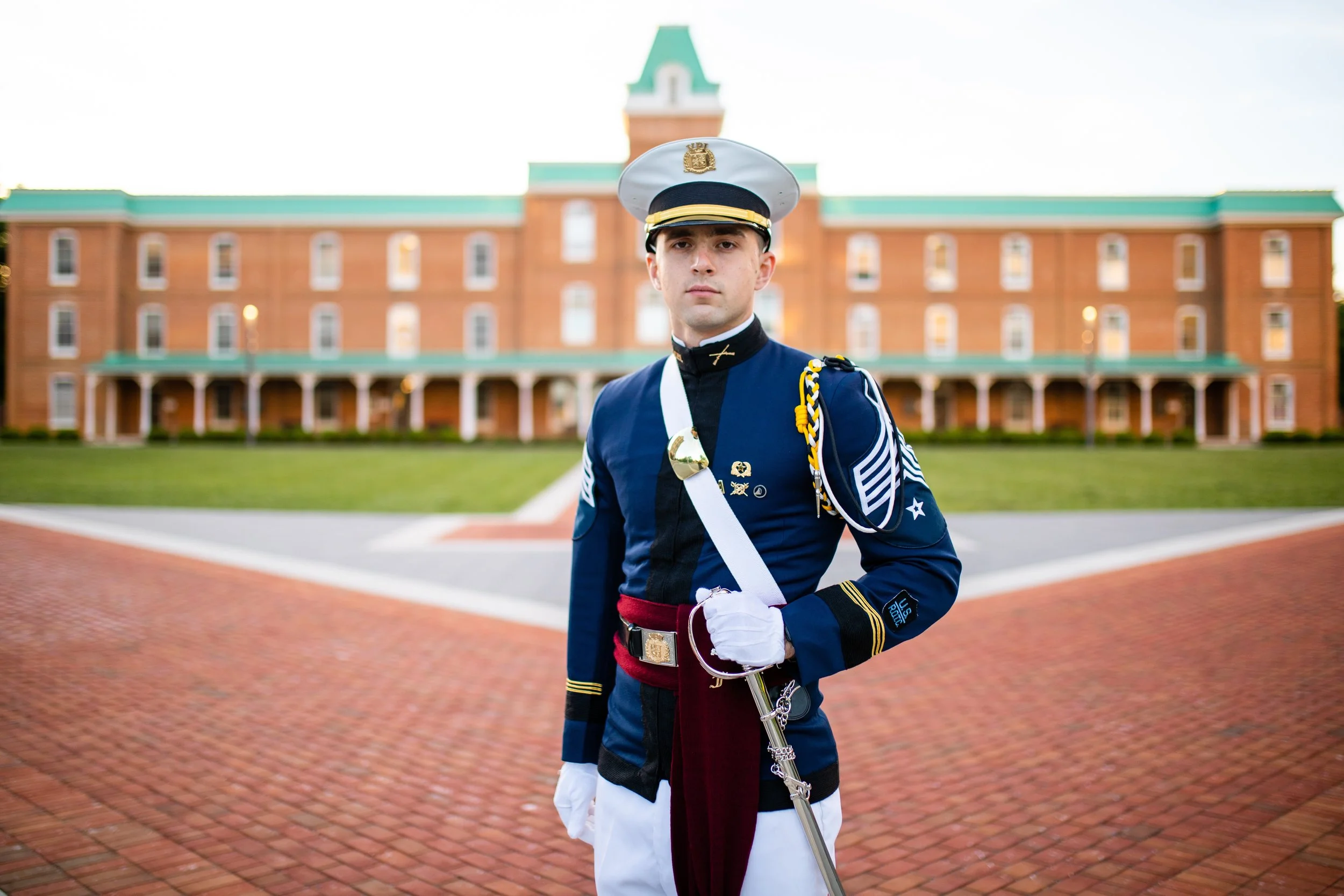 A young man in a military dress uniform standing in front of a brick building with green roofing, holding a sword.