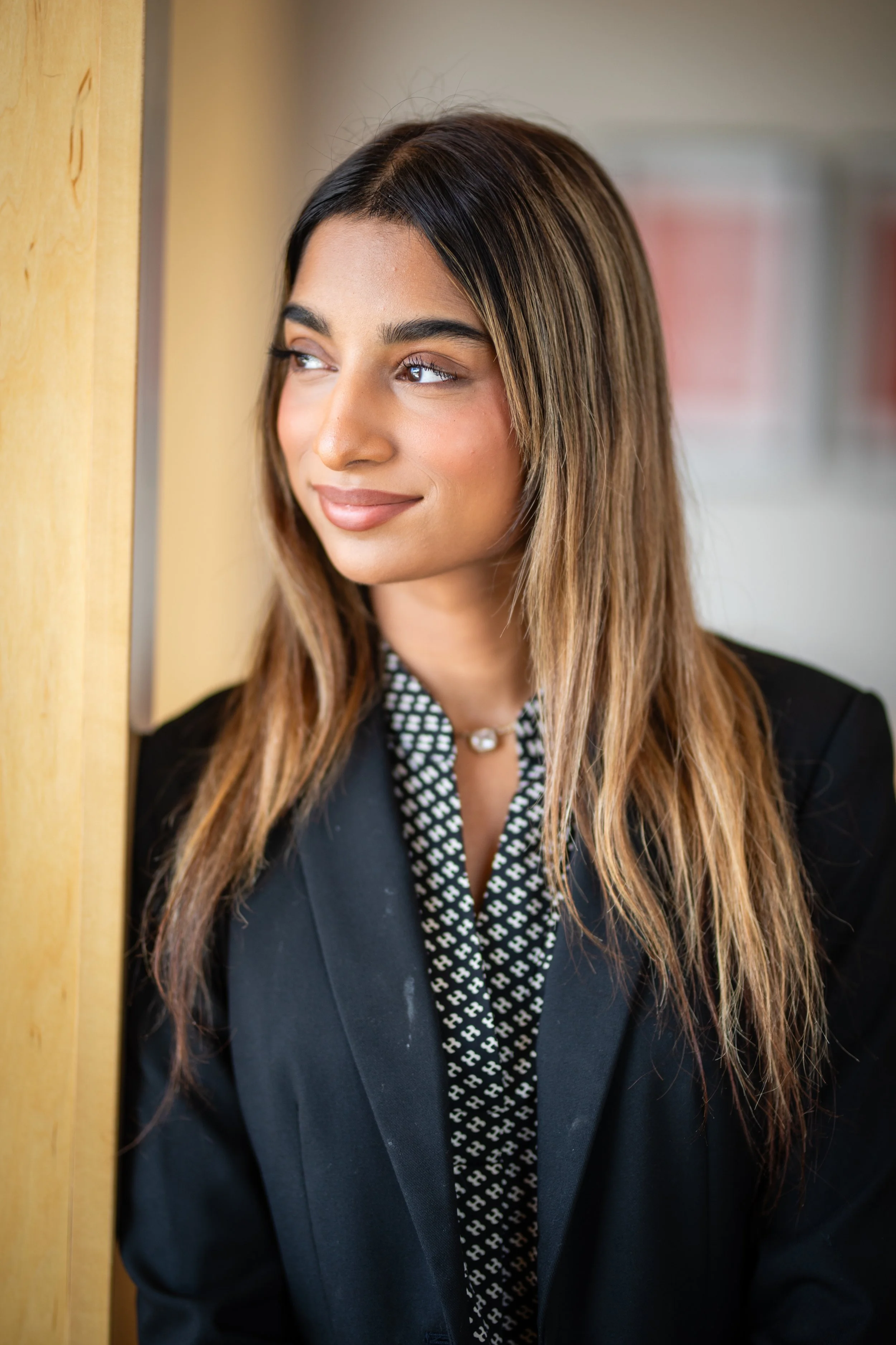 Portrait of a young woman with long brown hair looking out a window, wearing a black blazer, patterned blouse, and pearl necklace.