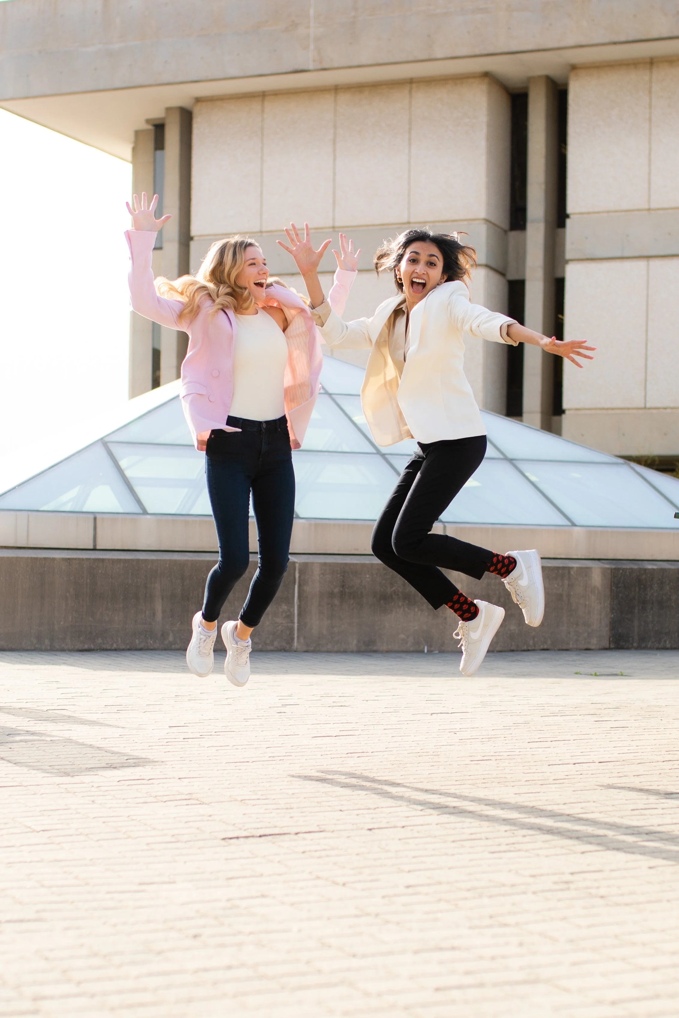 Two women jumping in the air outdoors in front of a modern building with glass and stone design, smiling and celebrating.