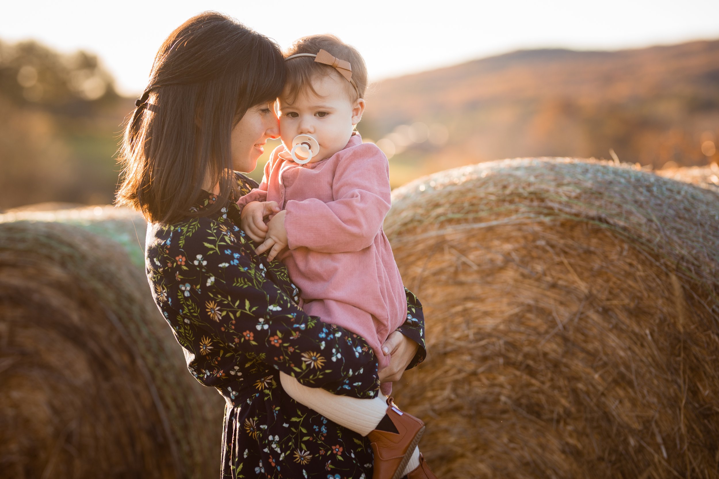 A woman holding a young girl with a pacifier in her mouth outdoors in front of large hay bales, during sunset.