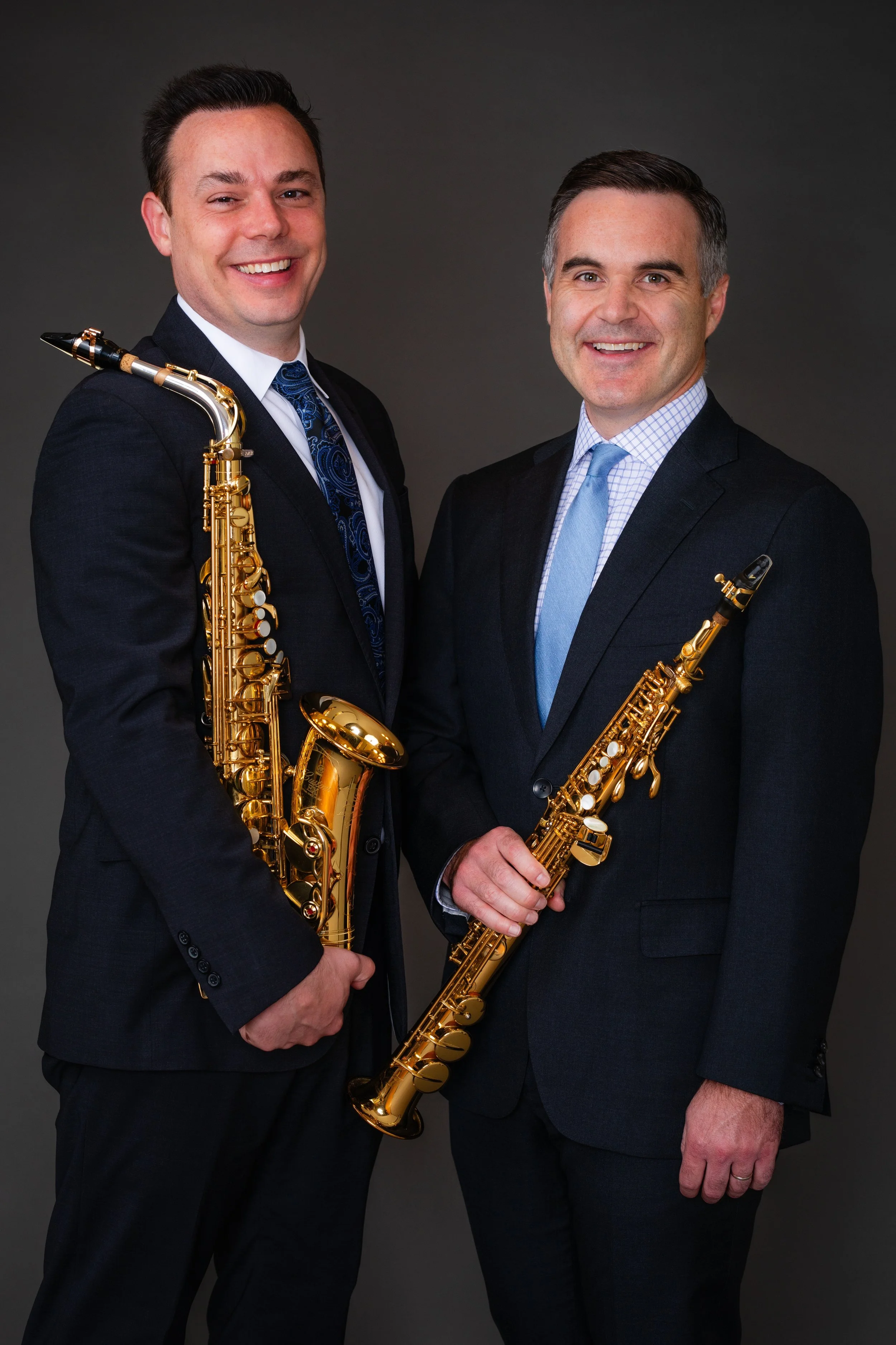 Two men dressed in suits holding saxophones and smiling at the camera against a dark background.