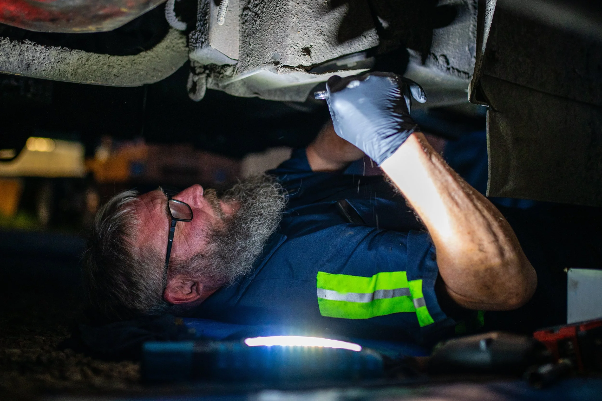 A maintenance worker with glasses, a beard, and wearing gloves, is lying on his side under a vehicle, inspecting or working on its underside in a workshop setting.