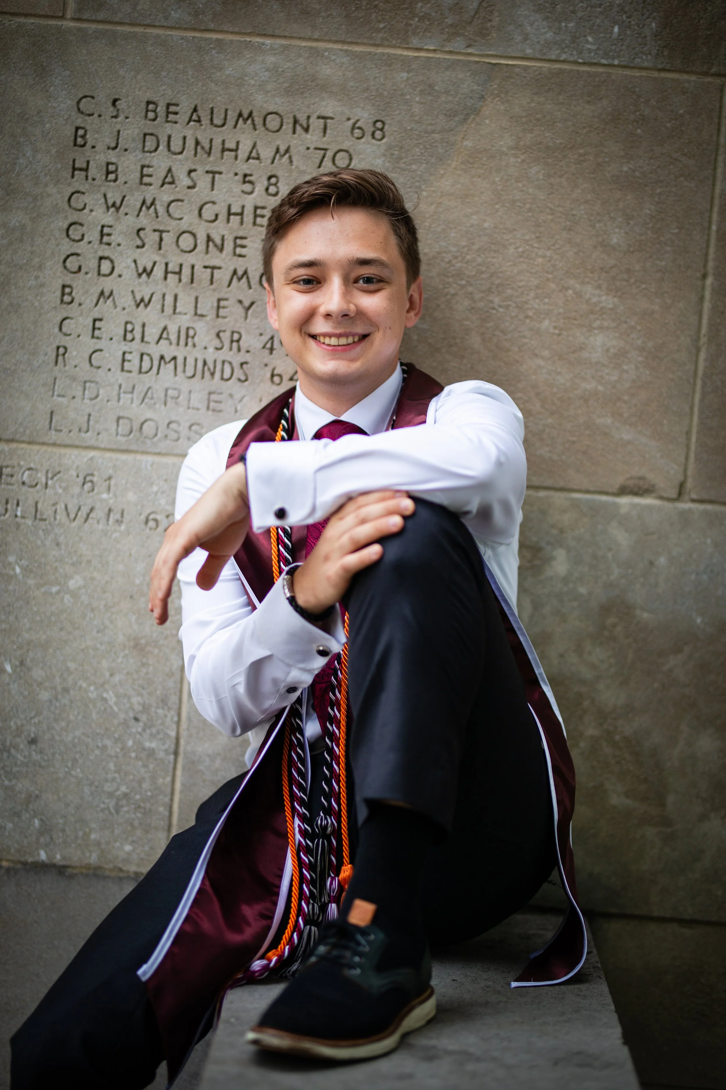 A young man sitting on a ledge in front of a stone wall with engraved names, dressed in a white shirt, maroon stole, black pants, and black shoes, smiling at the camera.