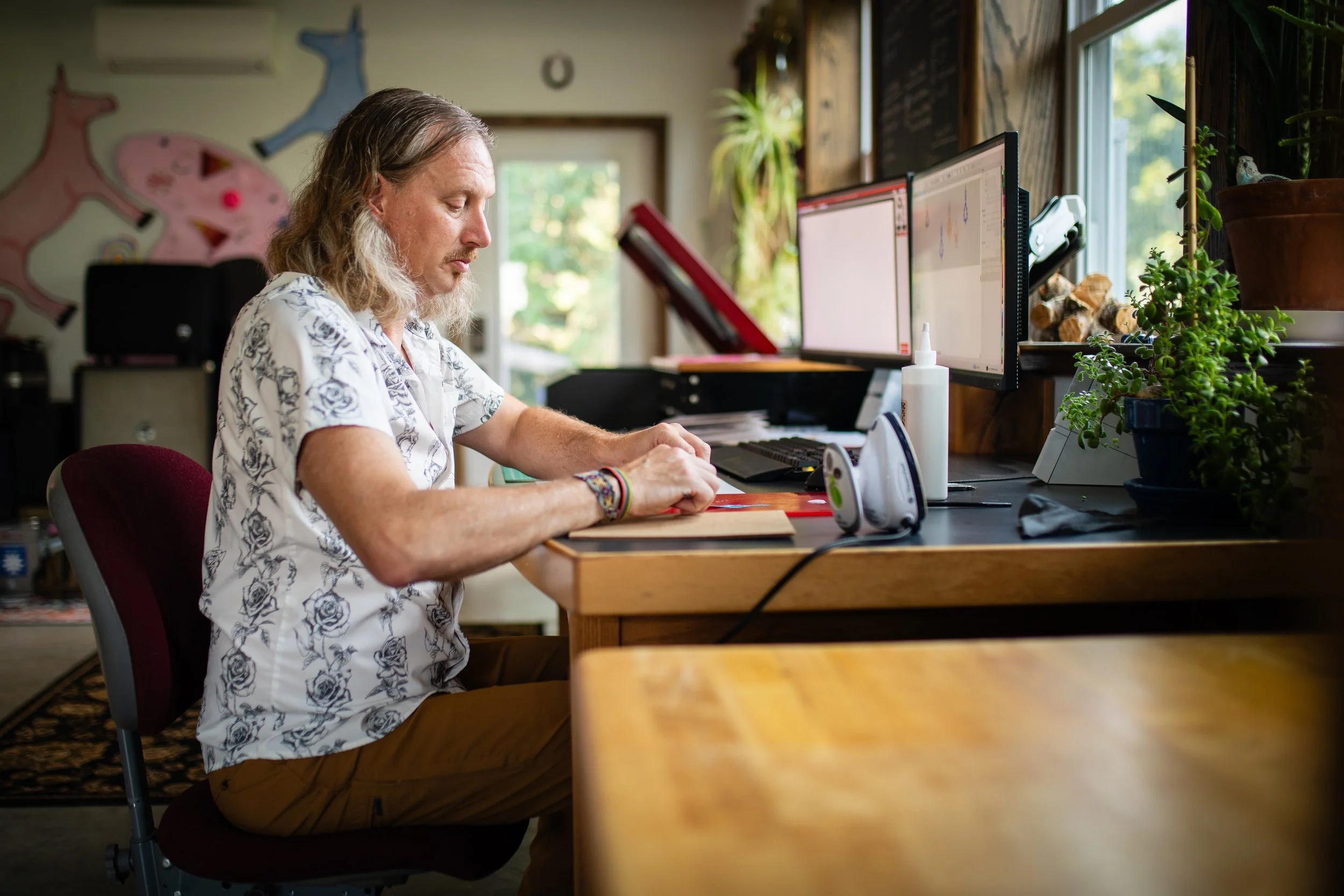 A man with long hair, wearing a white shirt with a floral pattern, sitting at a desk in a room with multiple computer monitors, plants, and decorations, writing or drawing on a piece of paper.
