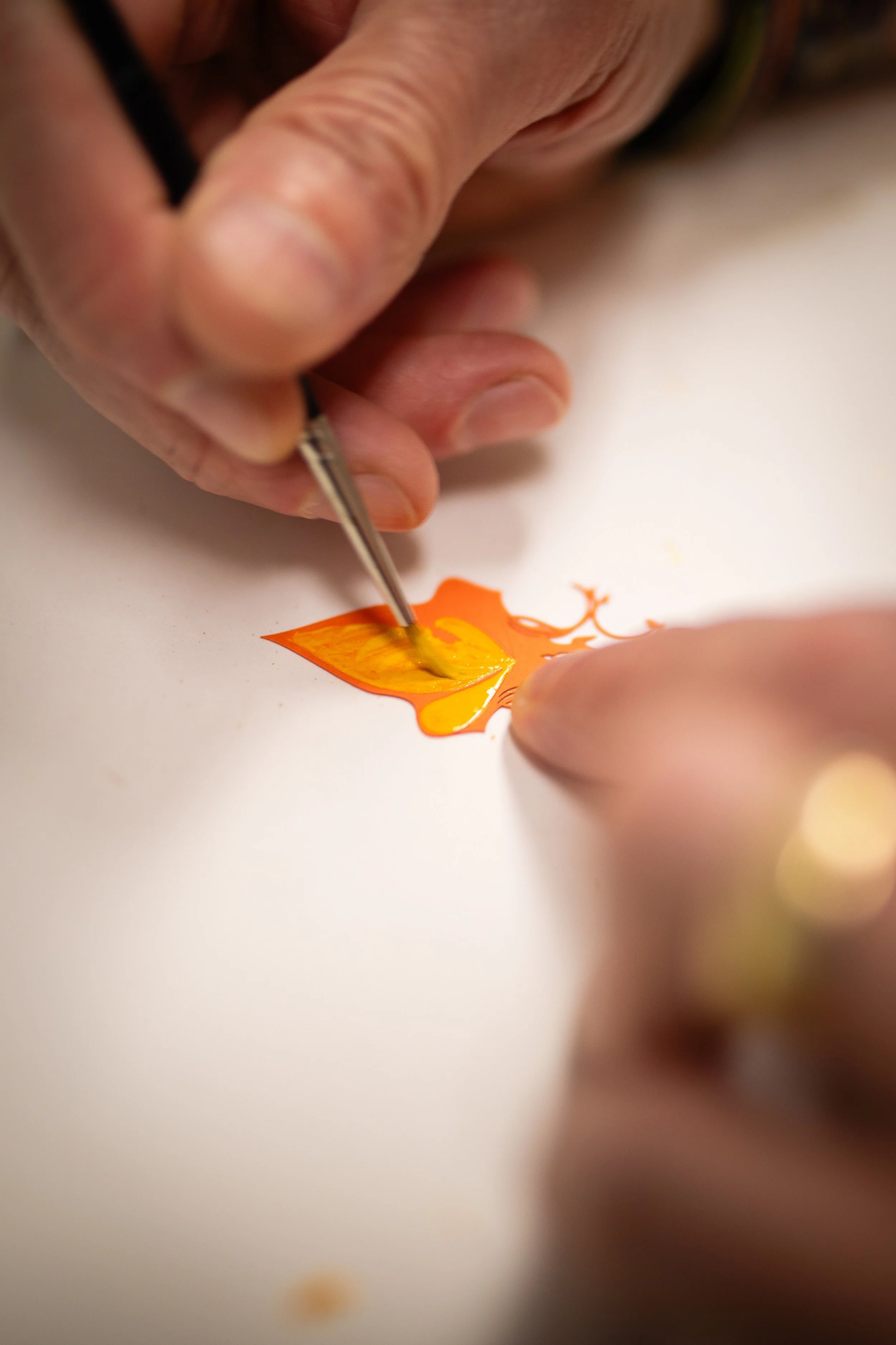 Close-up of an artist's hand using a fine brush to paint an orange and yellow design on a white surface.