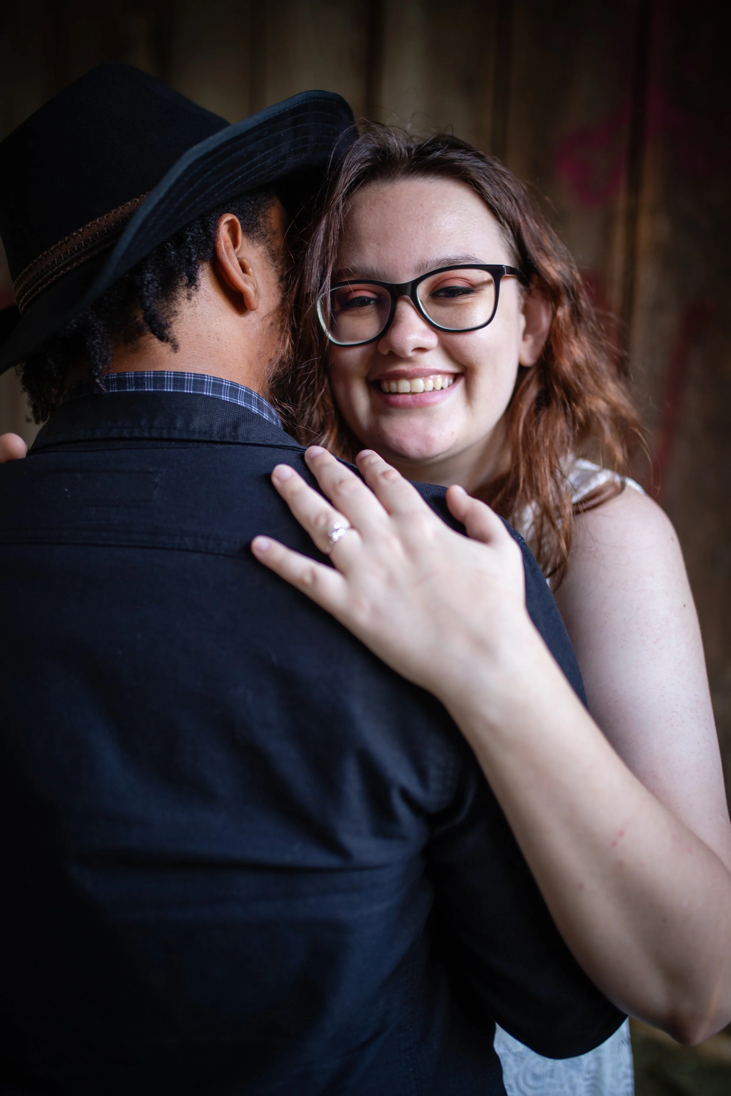 A smiling woman with glasses embraces a man wearing a black hat and dark clothing, indoors with a wooden wall background.