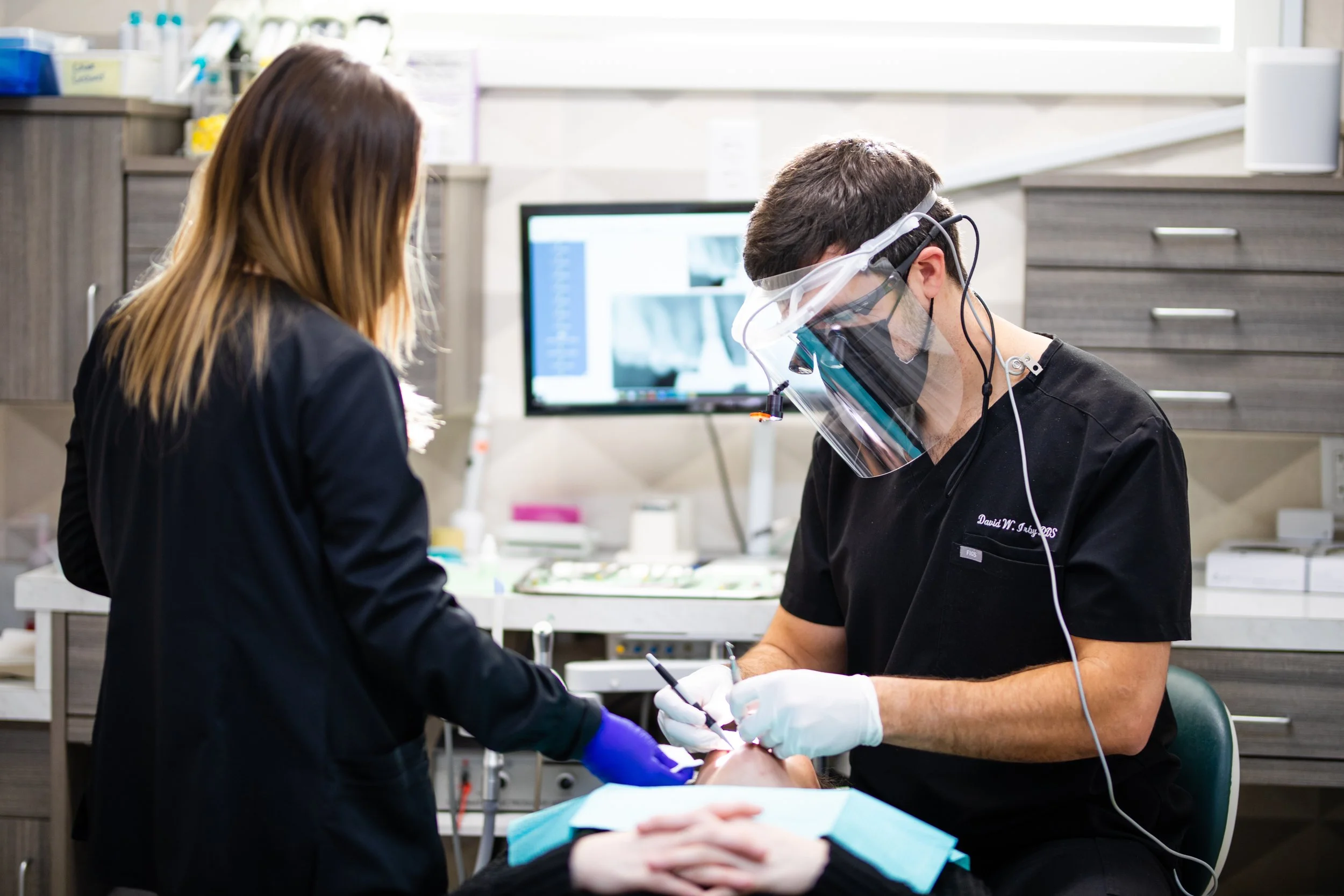 A dentist wearing a face shield and gloves performs a dental procedure on a patient in a dental clinic, with a dental assistant standing nearby and dental equipment visible.