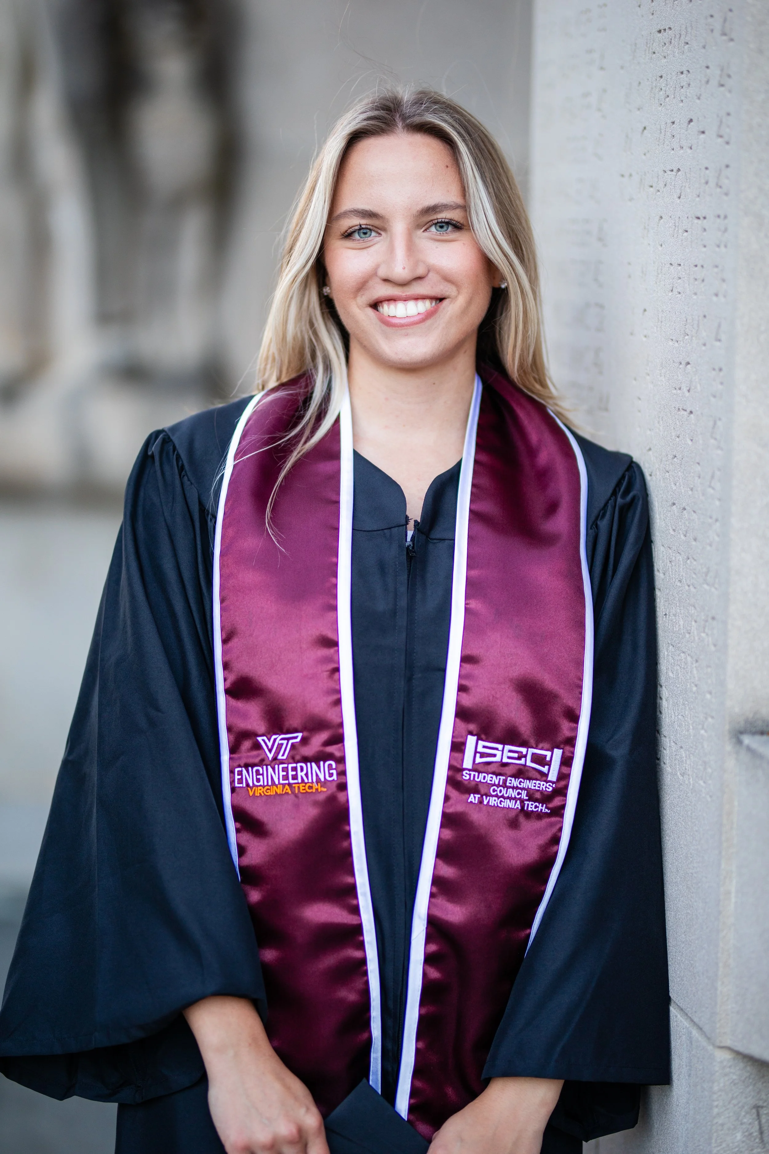 A young woman in a graduation gown and maroon stole smiling while standing against a stone wall.