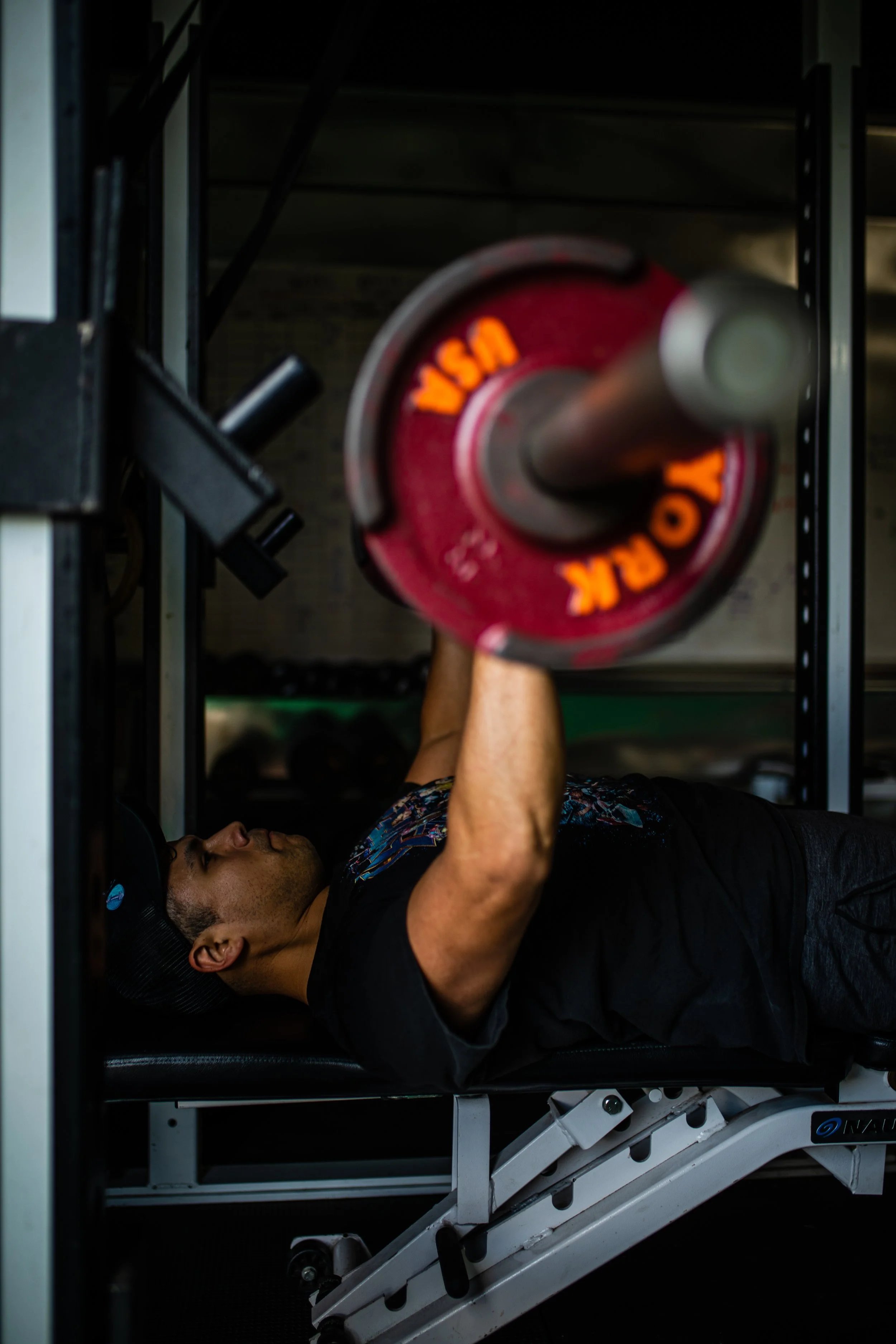 A man lying on a workout bench, lifting a barbell with red weight plates labeled 'YSA' during a strength training session in a gym.
