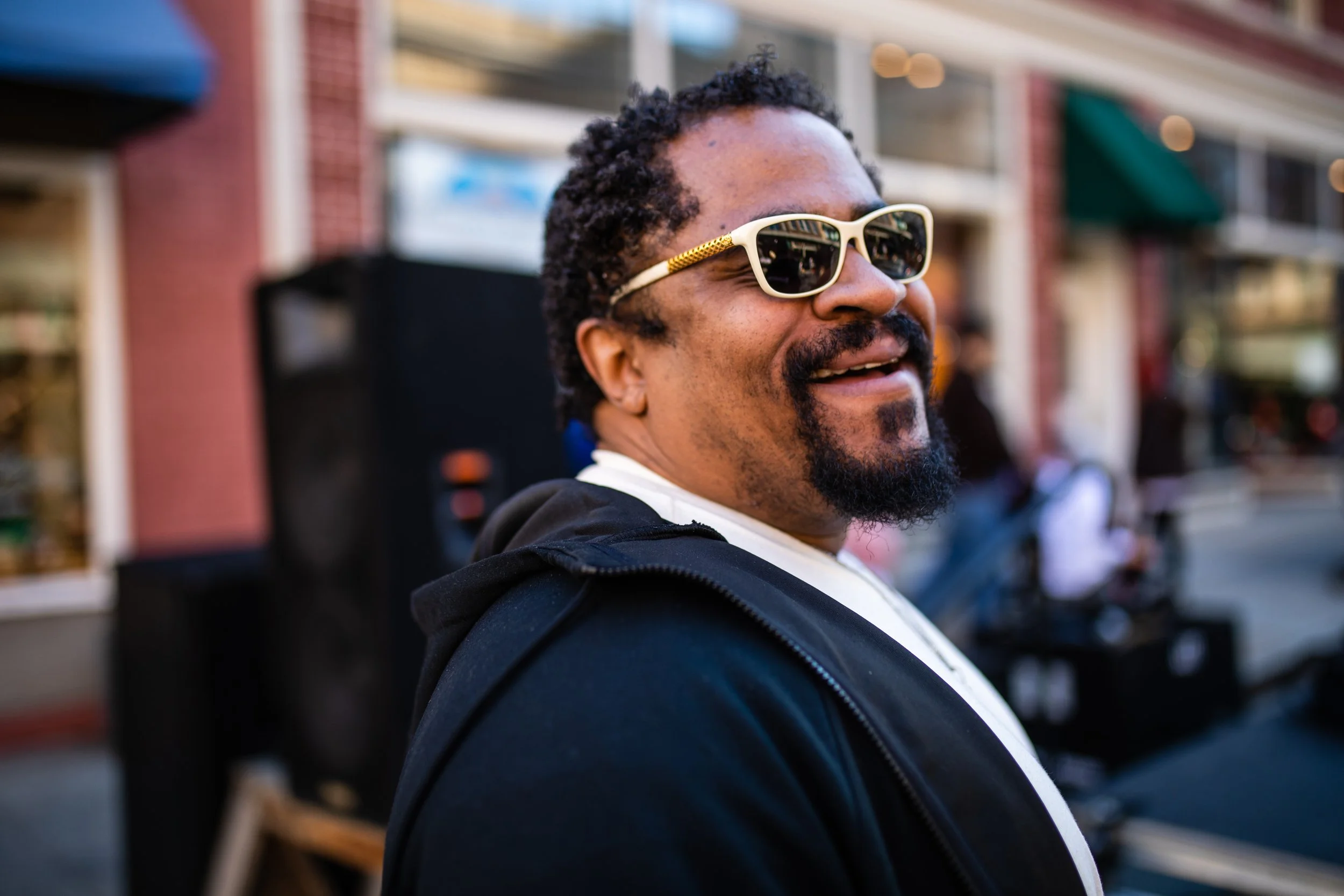 A man with a beard and curly hair wearing sunglasses and a black jacket smiling outdoors.