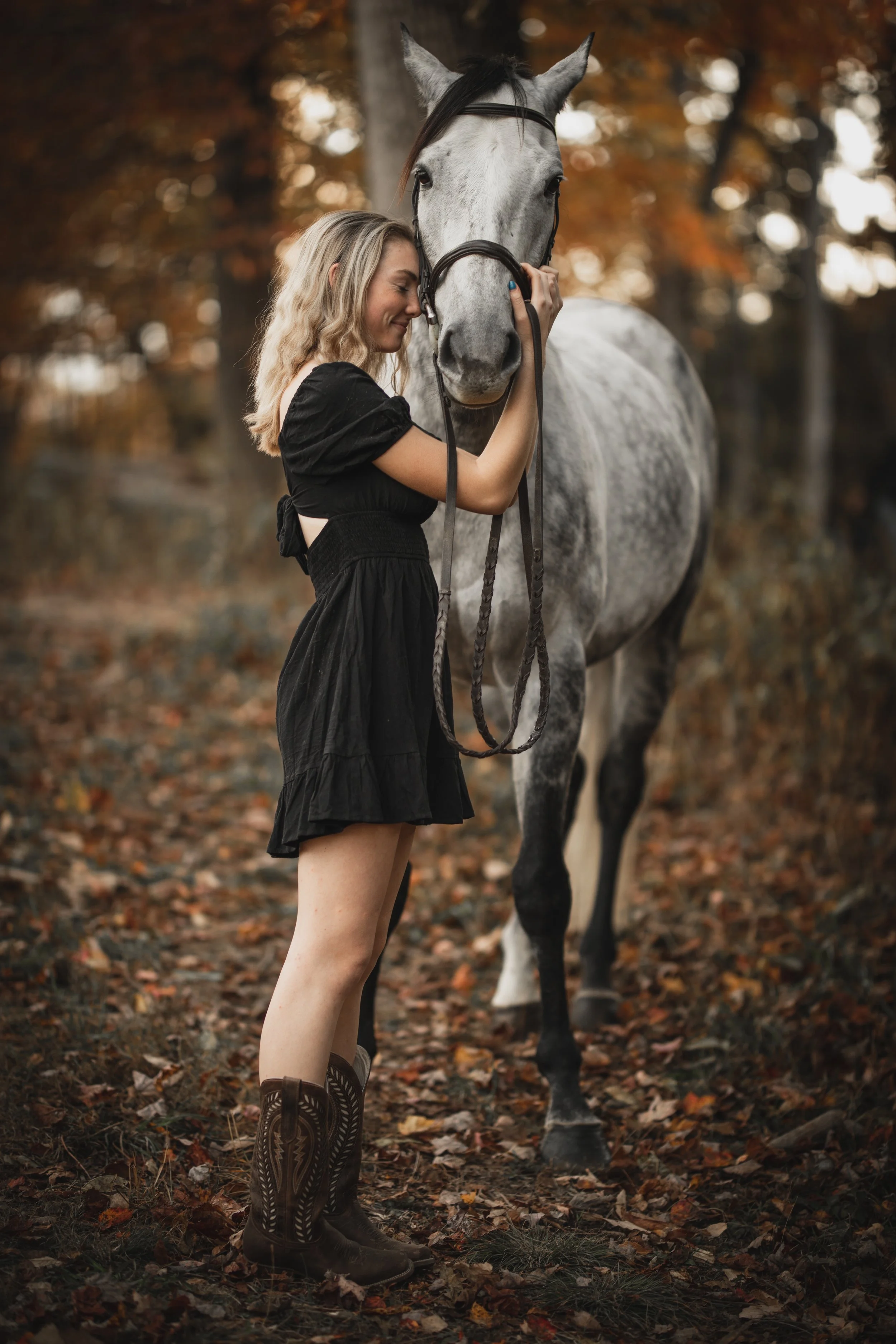 A woman hugging a gray horse in a forest with autumn leaves on the ground.