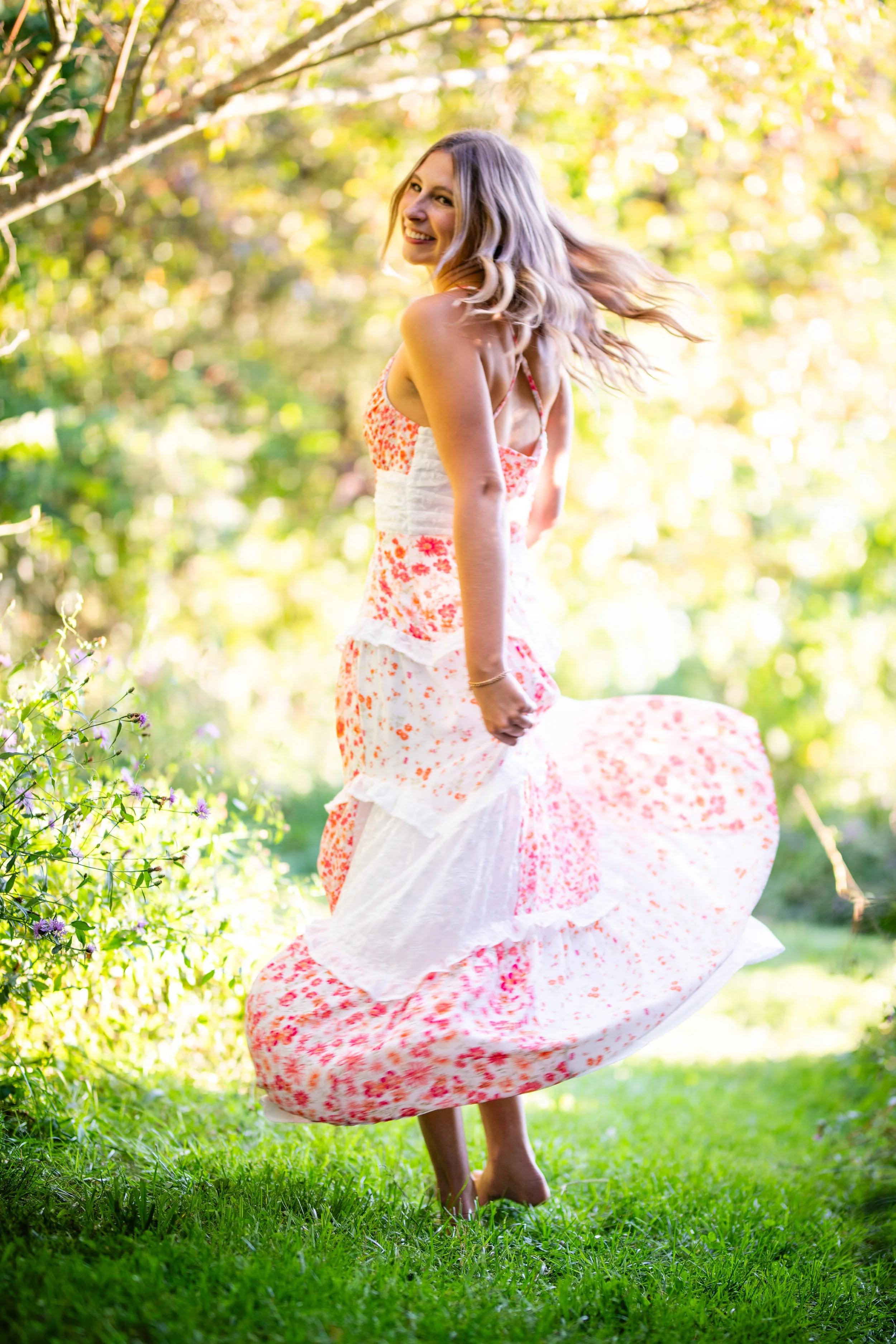 A smiling woman in a flowing floral dress twirling outdoors on grass, with green and yellow foliage in the background.