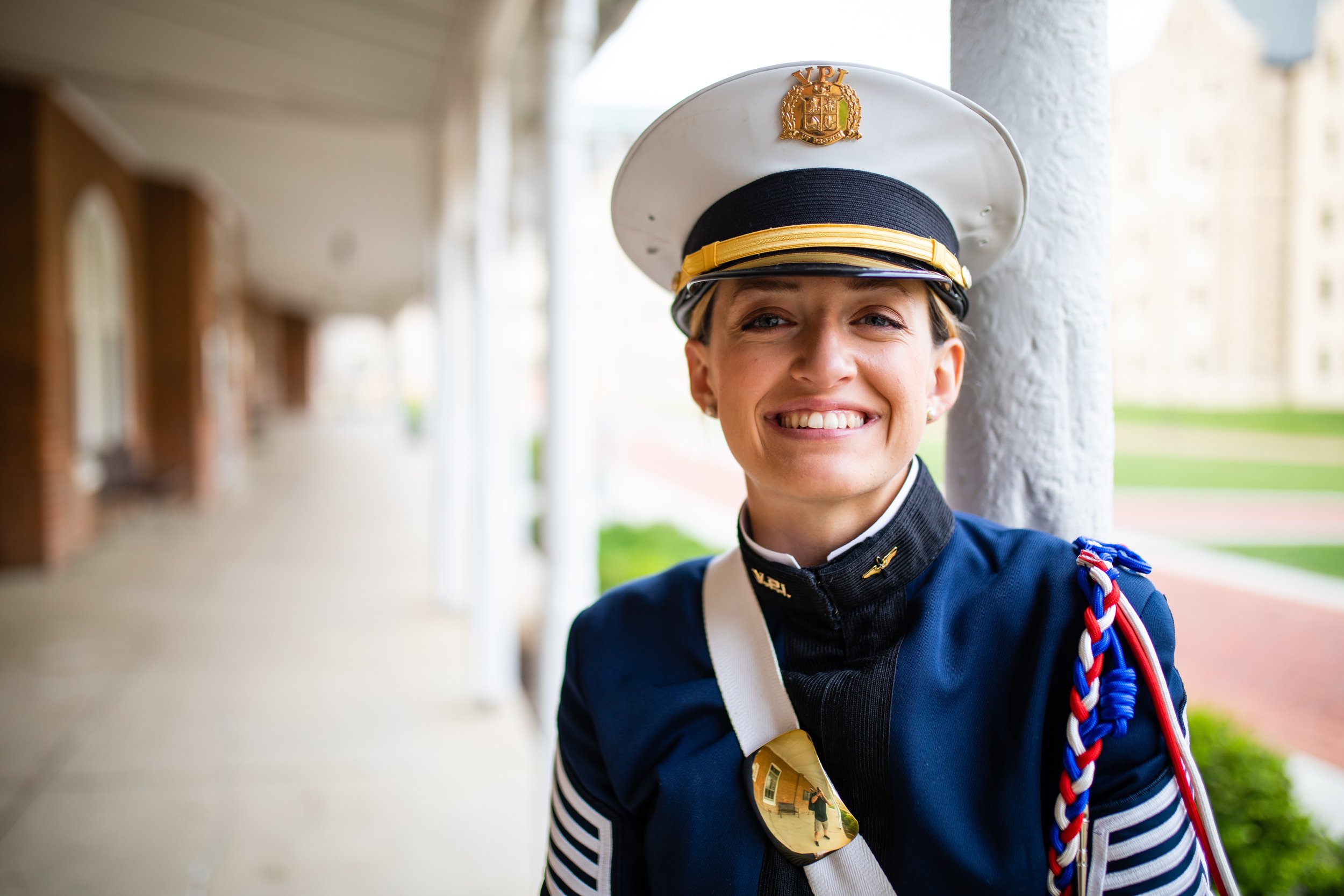 A woman in a military uniform smiling outdoors, wearing a white cap with a badge, sunglasses hanging on her uniform, and a decorated shoulder strap.