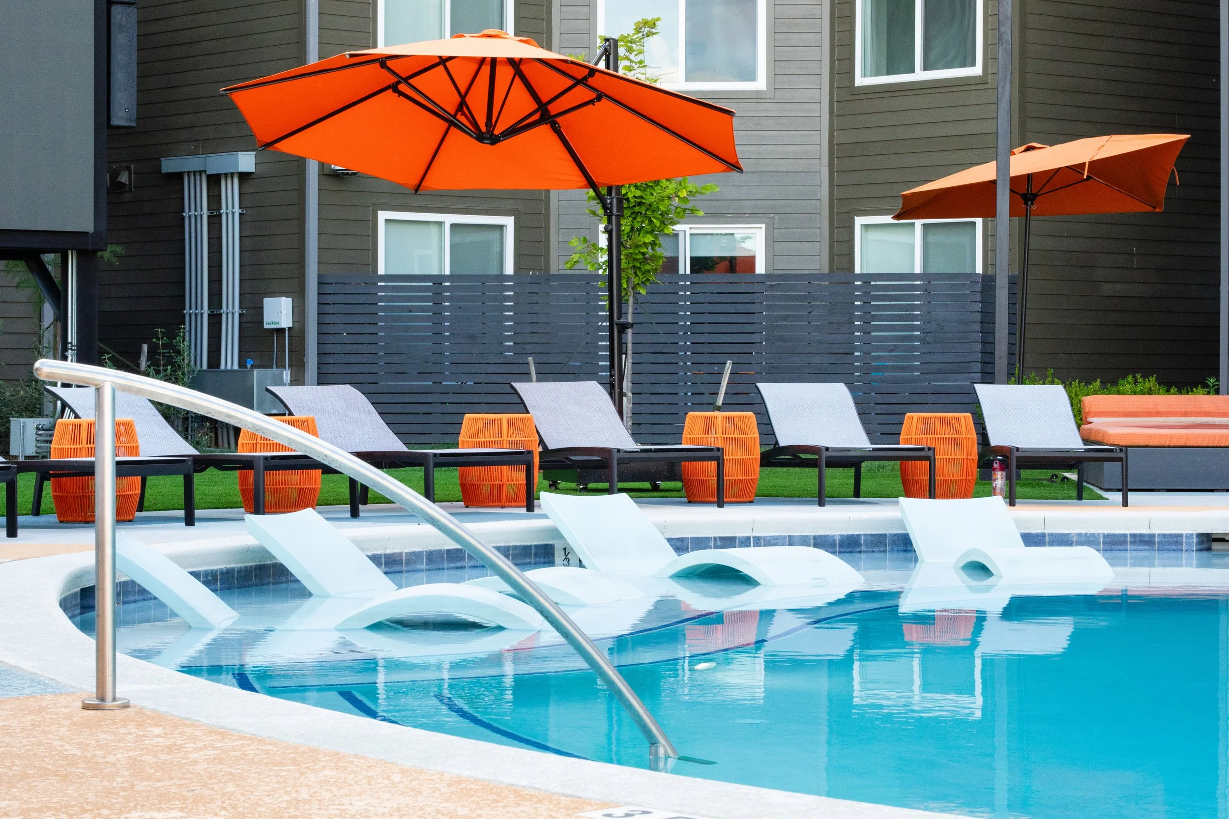 Artificial swimming pool with multiple water slides, orange umbrellas, lounge chairs, and a modern residential building in the background.