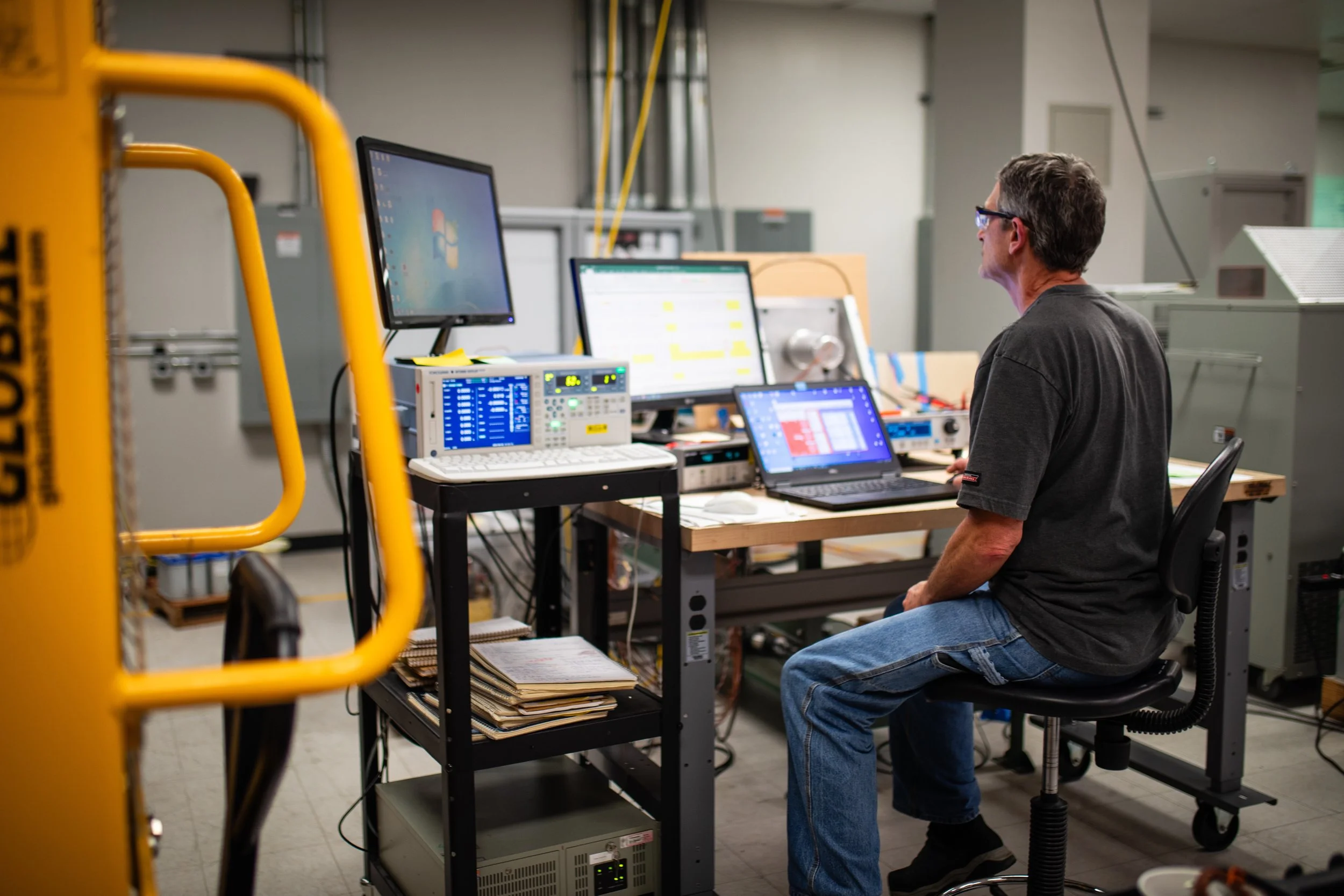 A man working at a desk with multiple computer monitors and electronic equipment in an industrial setting.