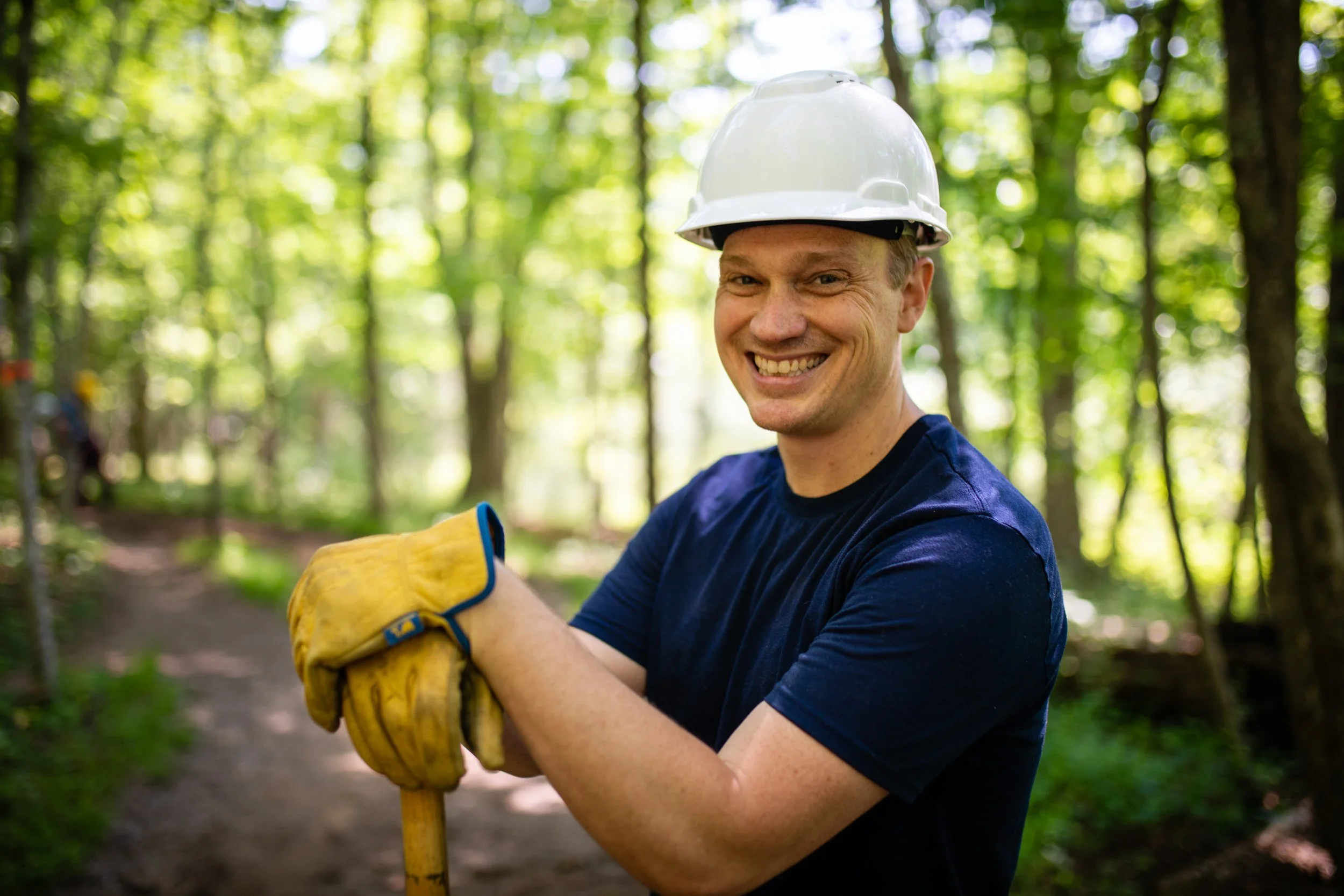 Smiling man wearing a white safety helmet and yellow work gloves, holding a shovel in a forest.