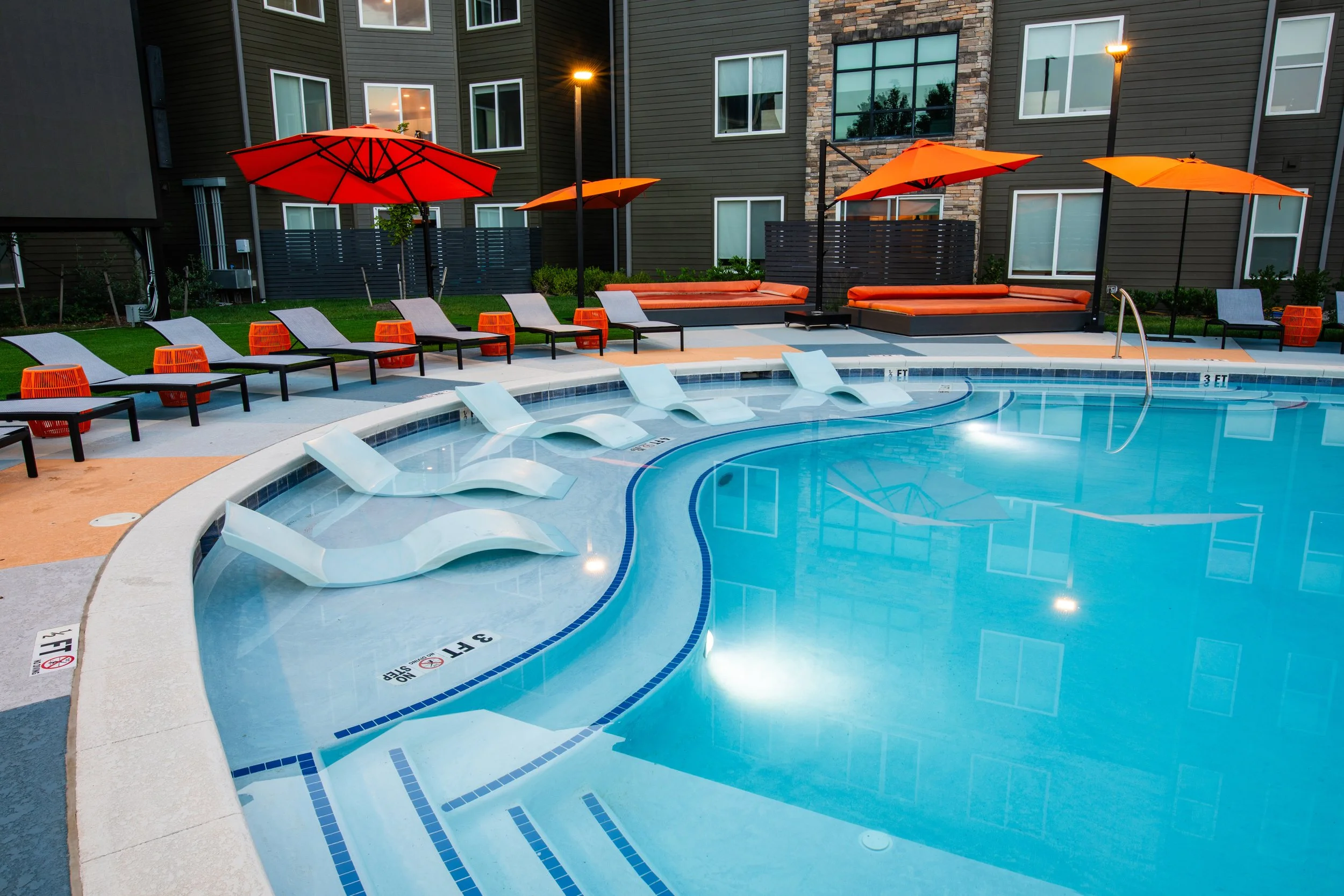 An outdoor swimming pool area with lounge chairs, orange umbrellas, and seating benches, set against a modern residential building at dusk.